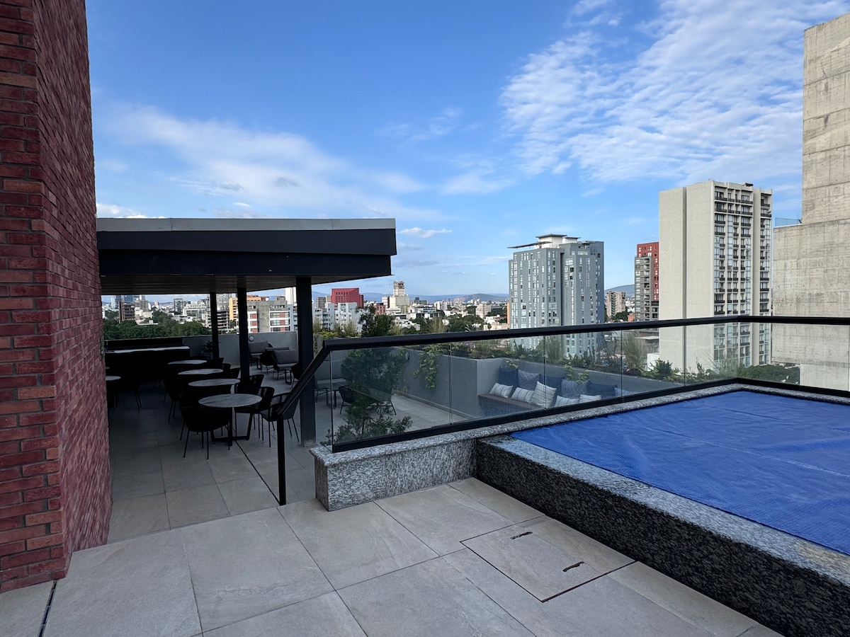 An outdoor terrace offers panoramic city views, complemented by comfortable seating areas. A sleek pool with a blue cover is visible, and a shaded dining area features several tables and chairs. The space is framed by modern architectural elements.
