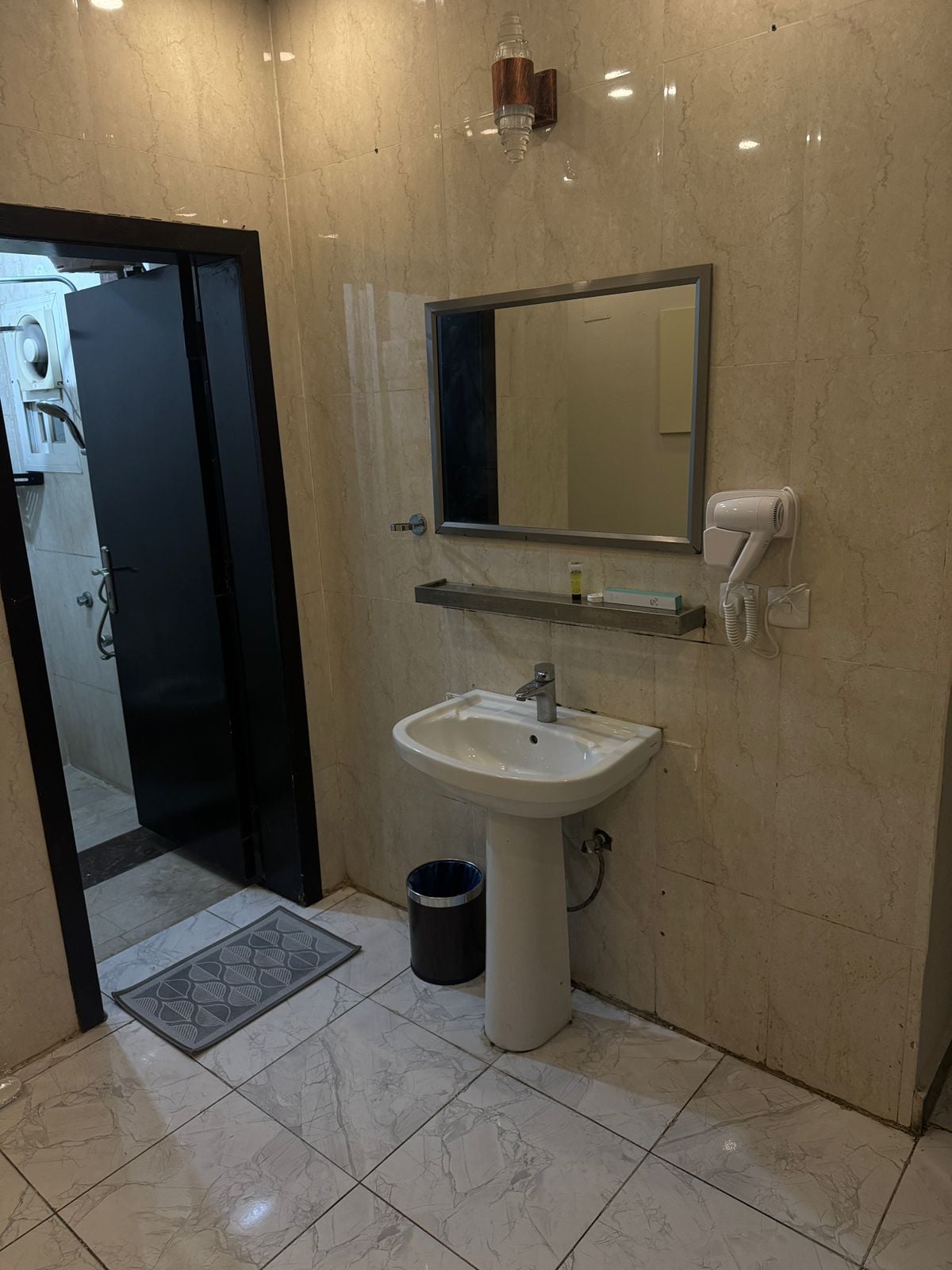 A well-lit bathroom is presented with beige tiled walls and a large mirror above a white pedestal sink. A wall-mounted hairdryer is visible beside the sink. A small shelf and a trash bin are also included. The entrance features a dark door leading to another area.
