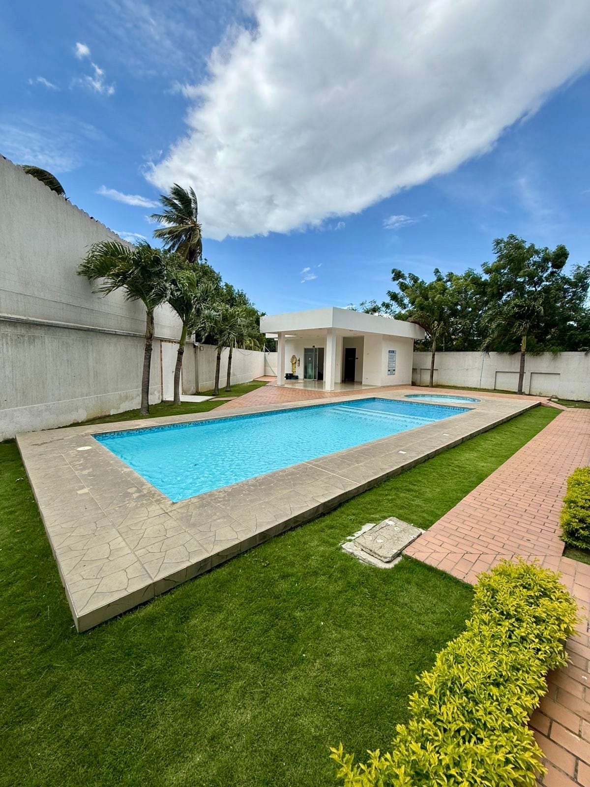 A serene outdoor area showcases a tranquil swimming pool surrounded by lush green grass. Palm trees frame the scene, and a modern structure is visible in the background. Sunlight reflects off the clear water, emphasizing the peaceful atmosphere of the space.
