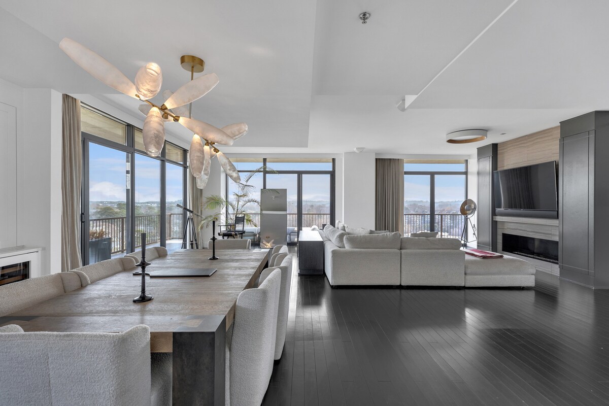 An open living space features a large dining table surrounded by textured chairs. A light fixture with multiple arms hangs above. Floor-to-ceiling windows provide expansive views, while a comfortable seating area with a soft sectional sofa is visible in the background.