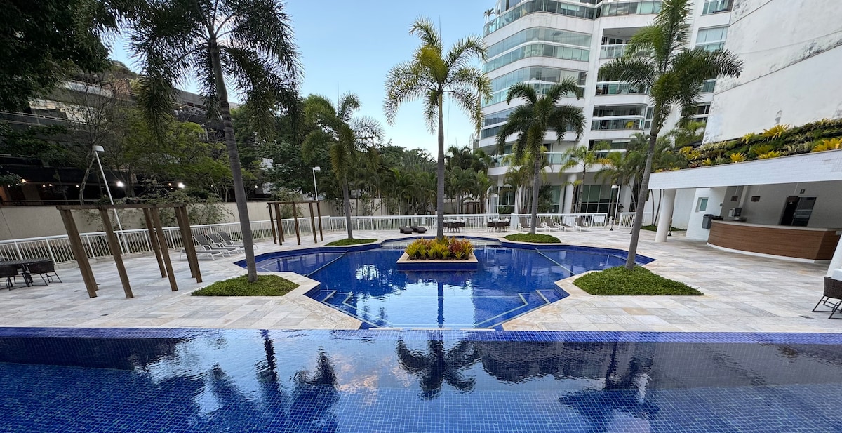 The image displays a serene swimming pool surrounded by lush palm trees. The clear blue water reflects the sky above, while well-maintained landscaping features vibrant flowers at the pool's center. Comfortable seating can be seen around the pool area, enhancing relaxation opportunities.