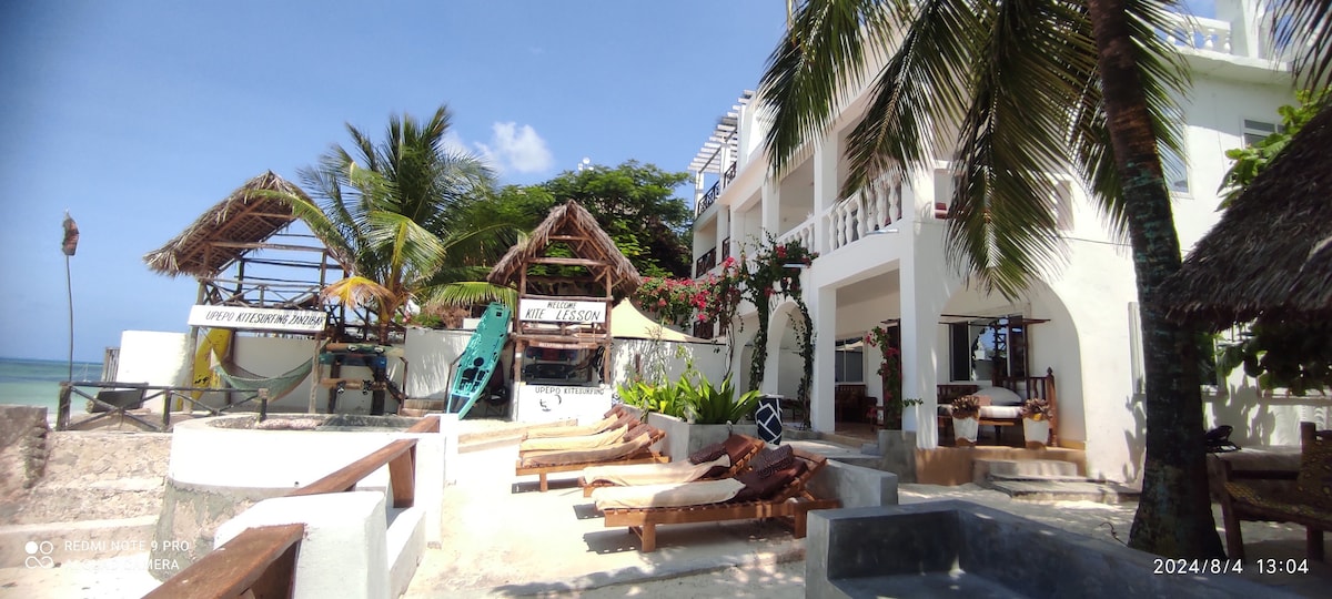 The beachfront area features a variety of seating options, including lounge chairs and shaded huts. Lush greenery surrounds the space, while the ocean is visible in the background. The building's white exterior blends with the coastal setting, providing an inviting atmosphere.