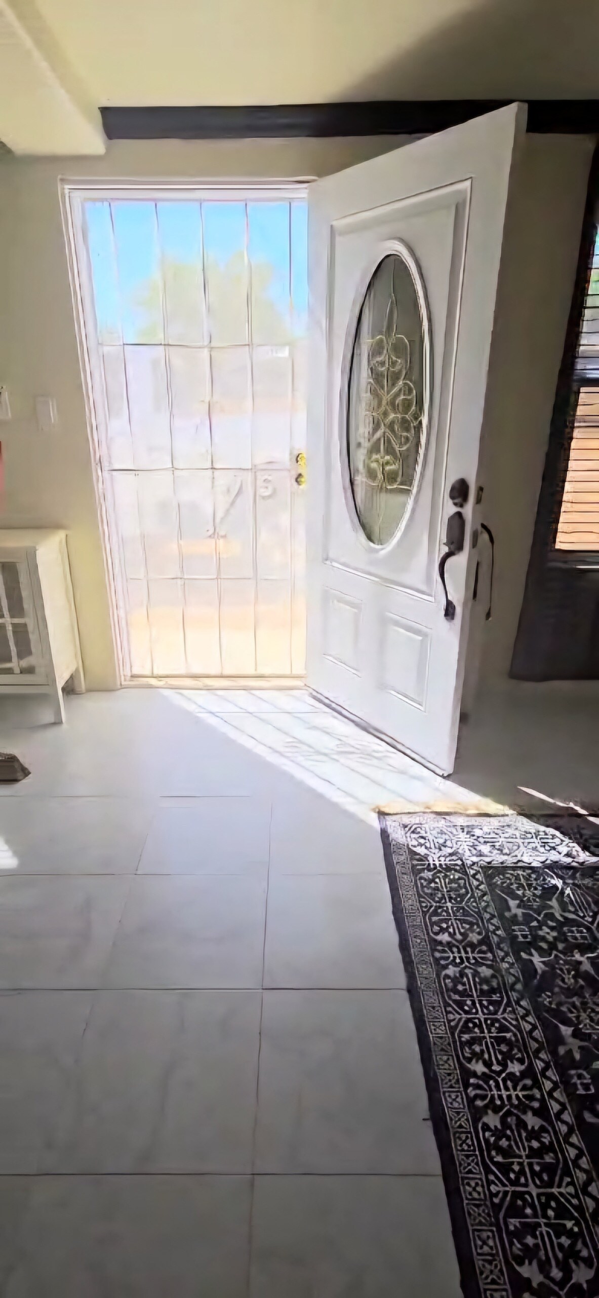 A welcoming entryway features a decorative glass door that allows natural light to filter in. Bright tiles line the floor, and a neutral-toned rug adds a soft touch. A small console table is positioned nearby, enhancing the open and airy feel of the space.