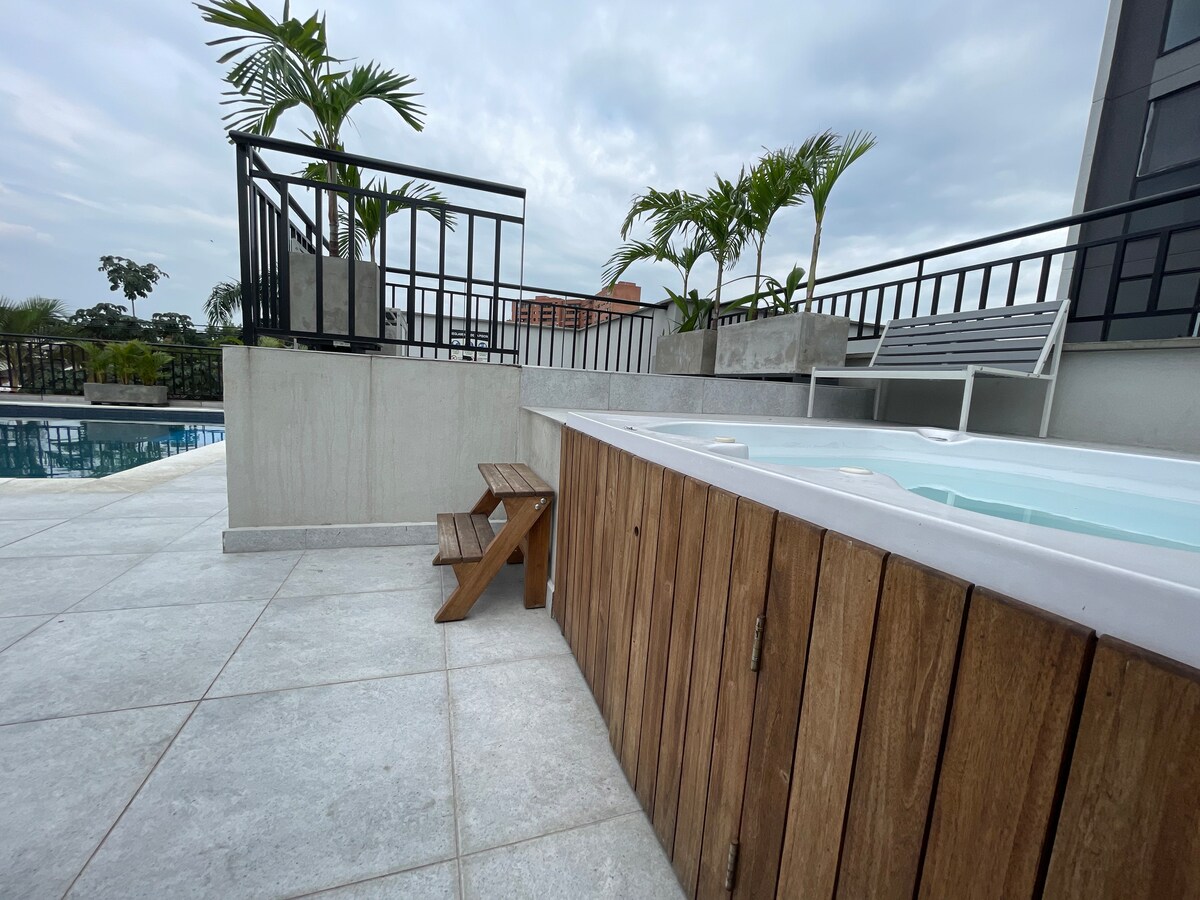 The rooftop area features a jacuzzi with wooden paneling, framed by modern railing and tropical plants. A small wooden step stool is positioned nearby. In the background, a distant swimming pool is visible, along with a spacious deck area designed for relaxation.