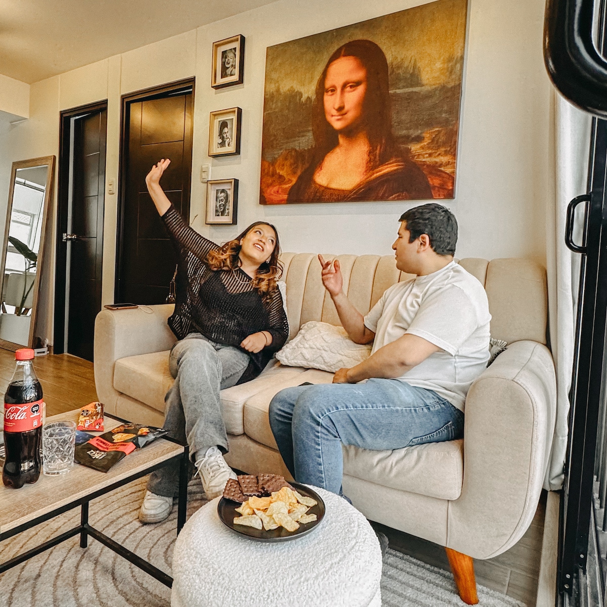 An inviting living area features a comfortable light-colored couch and a round coffee table adorned with snacks and beverages. A large artwork of the Mona Lisa hangs above, contributing to the room's artistic ambiance. Two guests are engaged in conversation, creating a welcoming atmosphere.