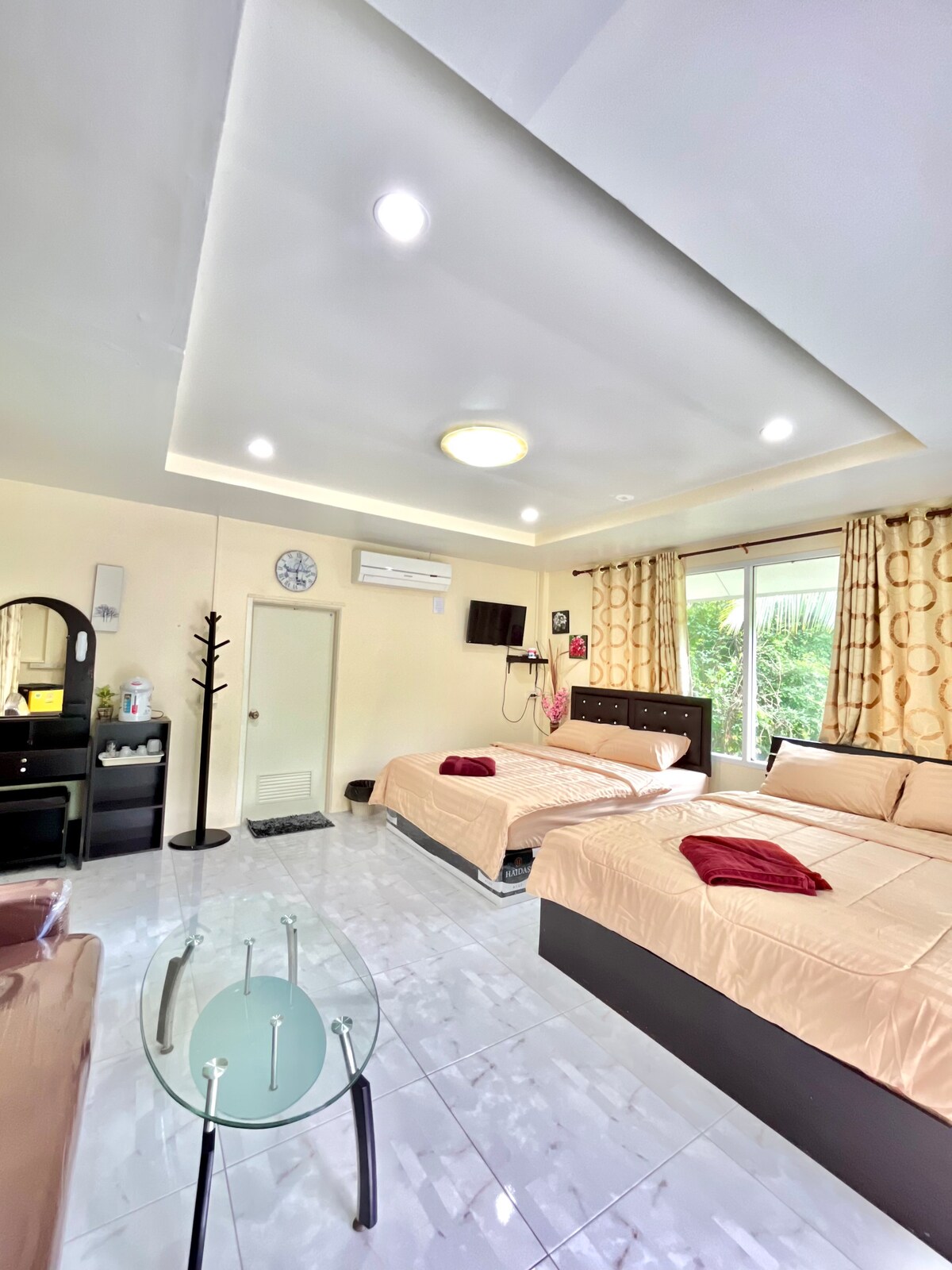 A spacious room is equipped with two queen-sized beds dressed in neutral bedding. A glass table is positioned within the area, while a makeup desk and entertainment unit are visible. Natural light flows in through a large window, framed with patterned curtains.