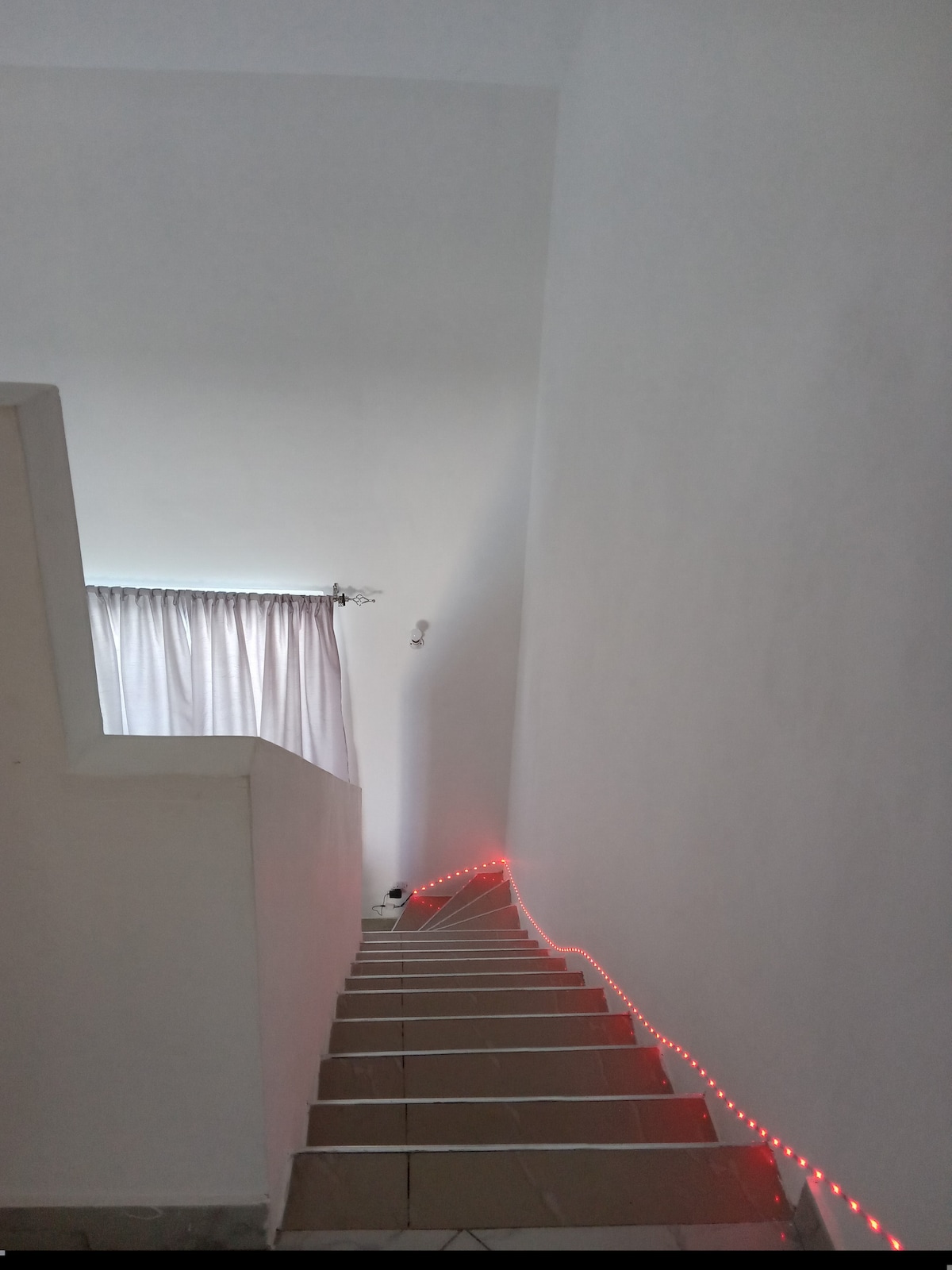 A flight of stairs is illuminated by a red light strip along the edge. The walls are painted white, and a window draped with light-colored curtains is visible at the top of the staircase, allowing natural light to brighten the space.