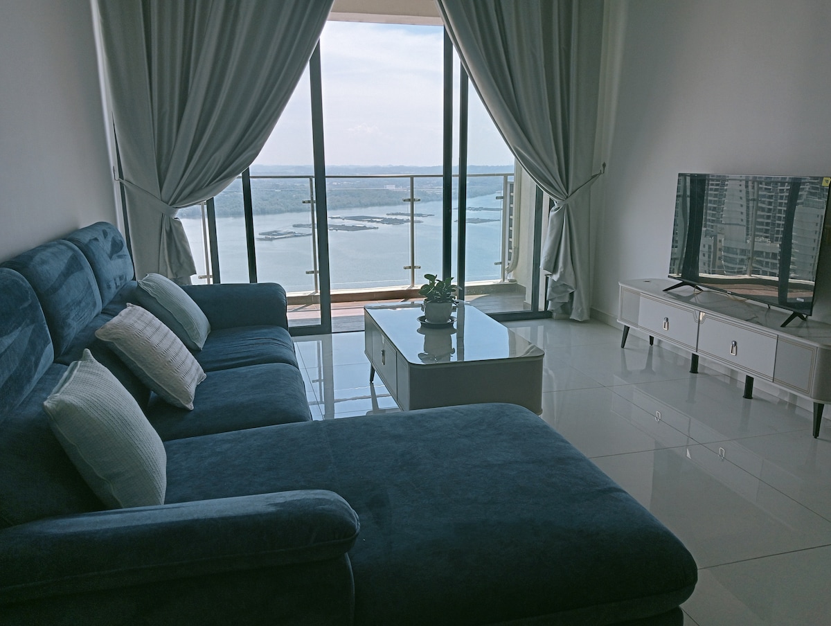 A spacious living area features a velvety blue sectional sofa arranged in front of a television. Large windows provide panoramic views of the water and distant skyline, with sheer curtains allowing natural light to fill the room. A simple coffee table is placed in the center.