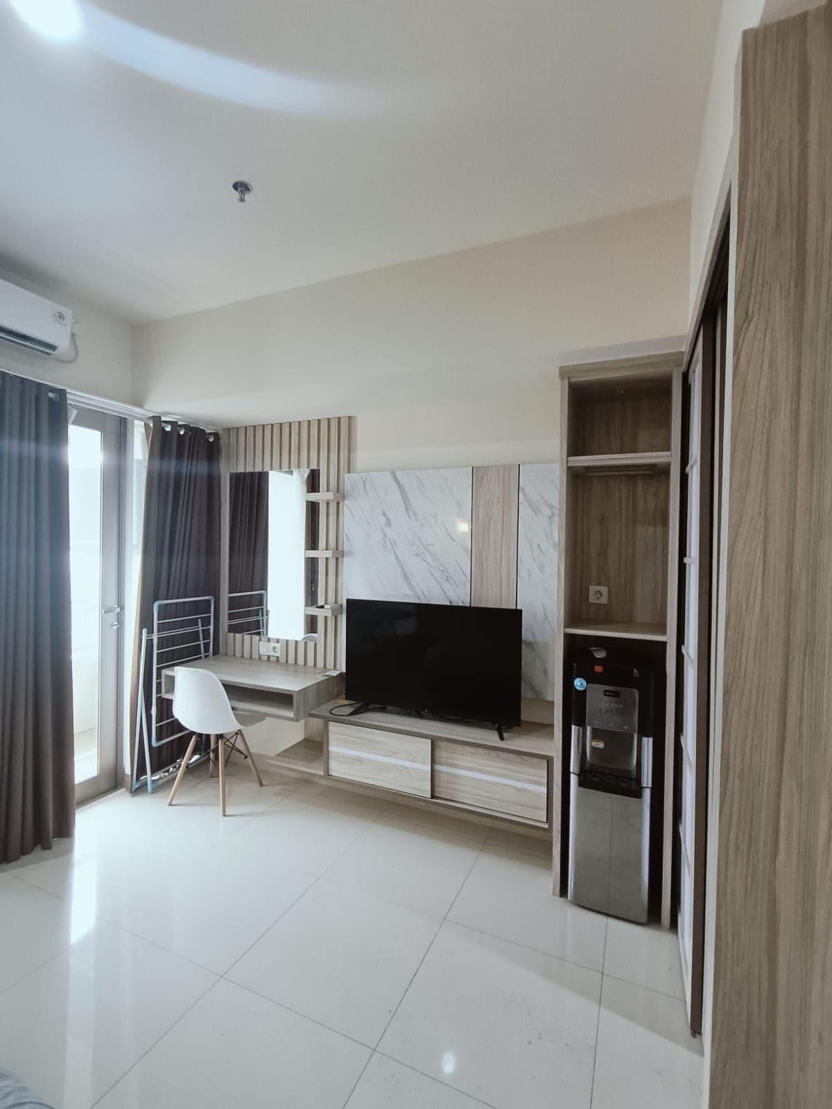 A modern room is showcased, featuring a large flat-screen TV on a wooden entertainment unit. A compact workspace with a chair is positioned near floor-to-ceiling windows draped with dark curtains. A mini-fridge is located beside the wardrobe, contributing to the room's functionality.