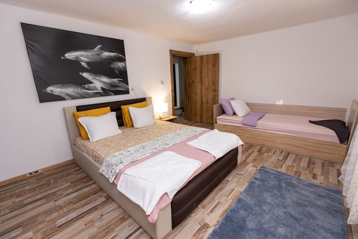 A spacious bedroom is presented with a large bed adorned with multiple pillows and layered bedding. A single bed is positioned alongside, featuring a simple design. The floor is covered with light wooden panels, complemented by a soft blue rug. Artwork of fish is displayed on the wall.