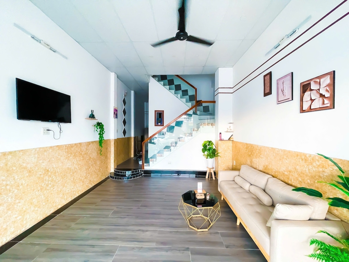 An open living area is featured, showcasing a light-colored sofa and a glass coffee table. A television is mounted on the wall opposite the sofa. A staircase with decorative tiles leads to upper levels, and greenery adds an inviting touch to the space.