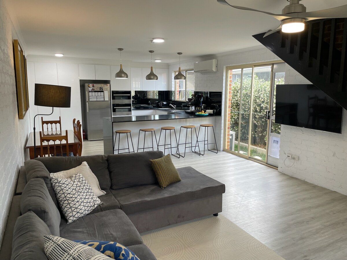 An open-plan living space features a spacious sectional sofa adorned with a decorative pillow. A dining table with wooden chairs is visible, and a sleek kitchen showcases modern appliances. Large glass doors lead to a private outdoor area with greenery, enhancing the airy atmosphere.