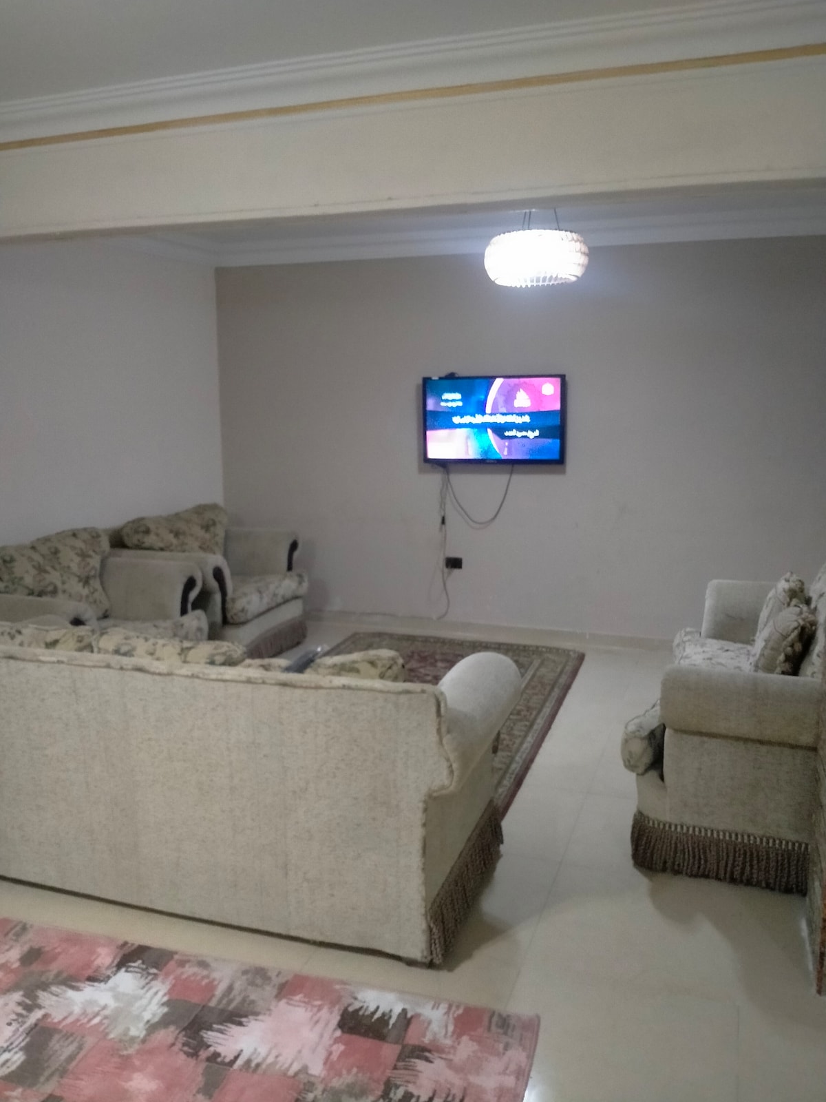 A comfortable living area features light-colored sofas arranged around a central area rug. A wall-mounted television is positioned in front of the seating, while a decorative light fixture hangs from the ceiling, illuminating the space.