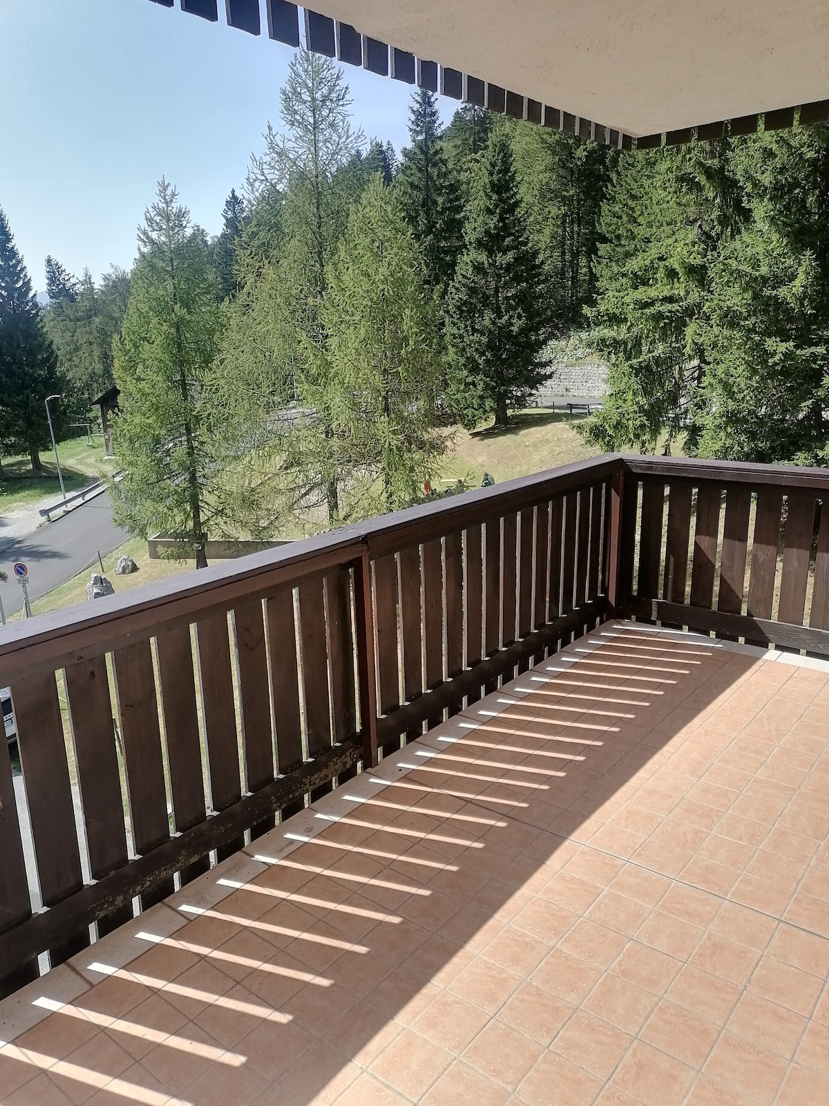 A wooden balcony is presented with a flat tile floor, offering views of a tree-lined landscape. Sunlight casts elongated shadows through the wooden railing, creating a warm and inviting space for relaxation in nature.