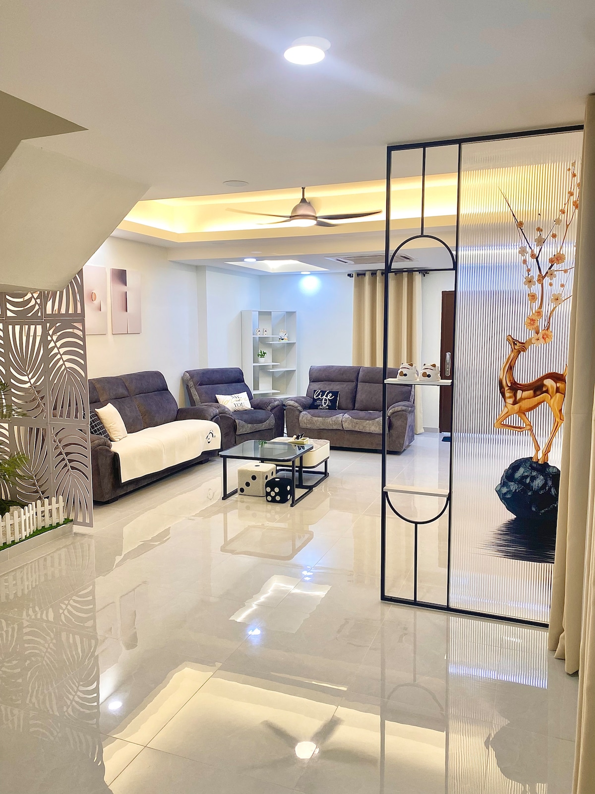 A modern living area features comfortable gray sofas, an accent chair, and a coffee table. Natural light illuminates the space, highlighting the glossy tiled flooring. A decorative screen separates the area from the adjacent room, adding visual interest with an art piece of a flower arrangement.