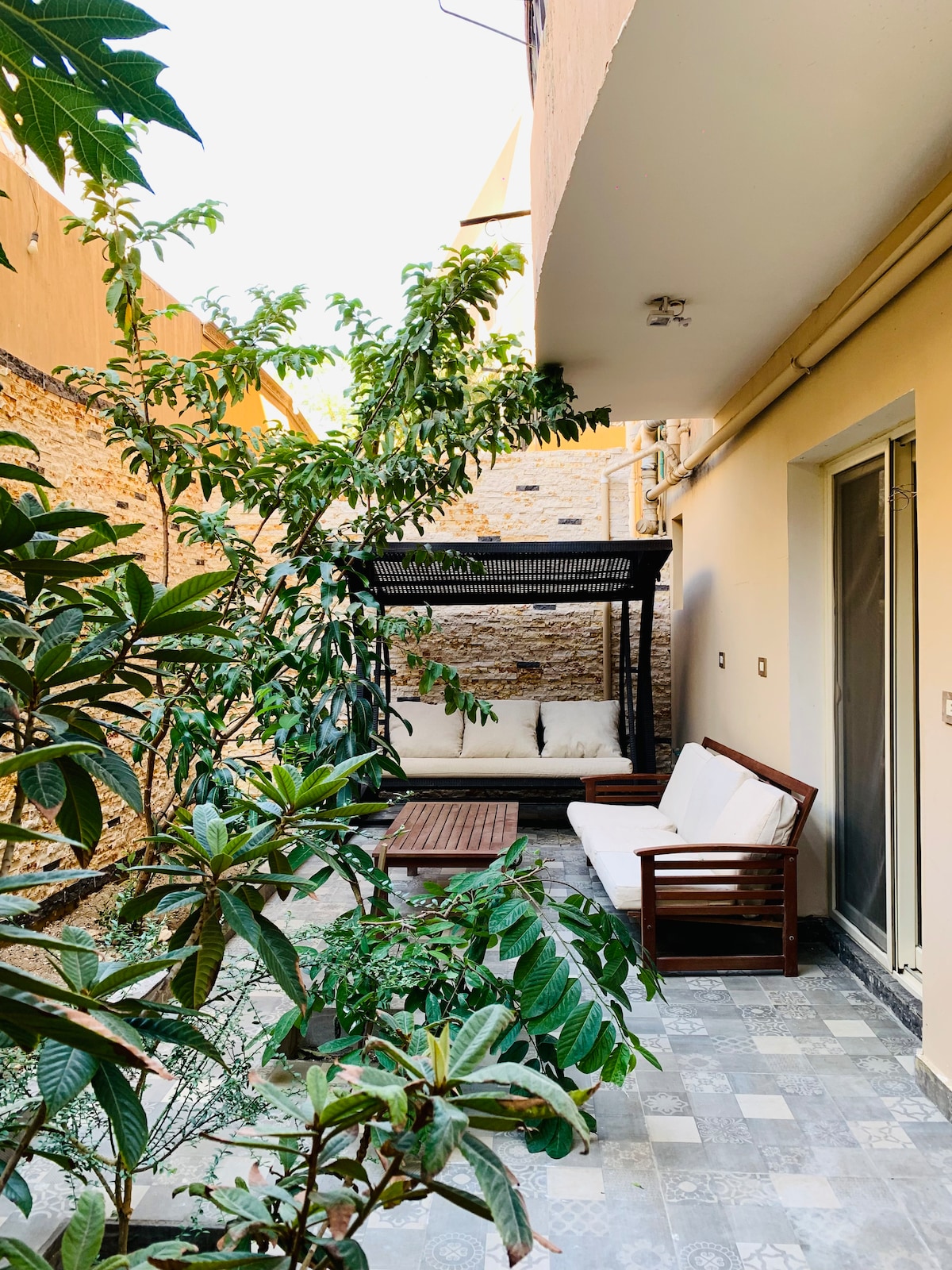 A private outdoor area features a wooden seating area with a cozy sofa, surrounded by lush greenery. A patterned stone floor adds to the natural ambiance. Soft, diffused light filters through the nearby plants, enhancing the serene environment.