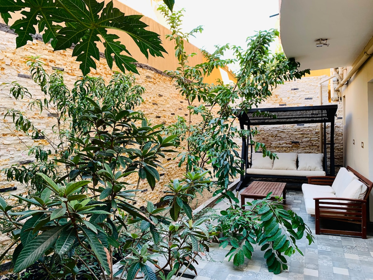 An outdoor space is surrounded by lush greenery, featuring various plants and trees. A comfortable seating area includes a wooden bench beneath a shaded pergola. Natural stone walls create a rustic backdrop, while patterned tiles cover the ground.