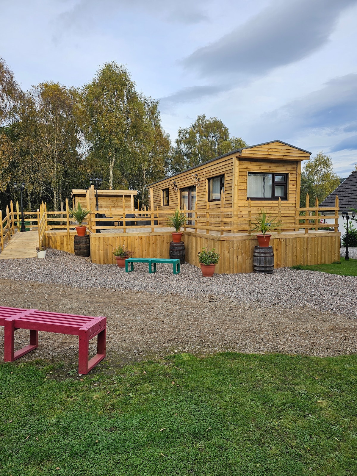 A wooden cabin designed for family stays is surrounded by a wooden deck and railings. The outdoor area features several potted plants and a gravel pathway, with benches offering seating. Natural surroundings provide a peaceful backdrop with trees in the distance.