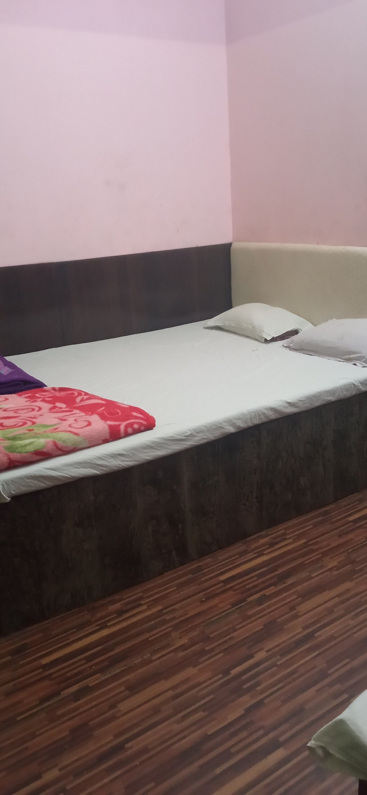 A sleeping area is presented with a spacious bed featuring a fitted mattress and several pillows. Two colorful blankets are arranged at the foot of the bed. The walls are painted in soft pink, while the floor has a warm wooden finish.