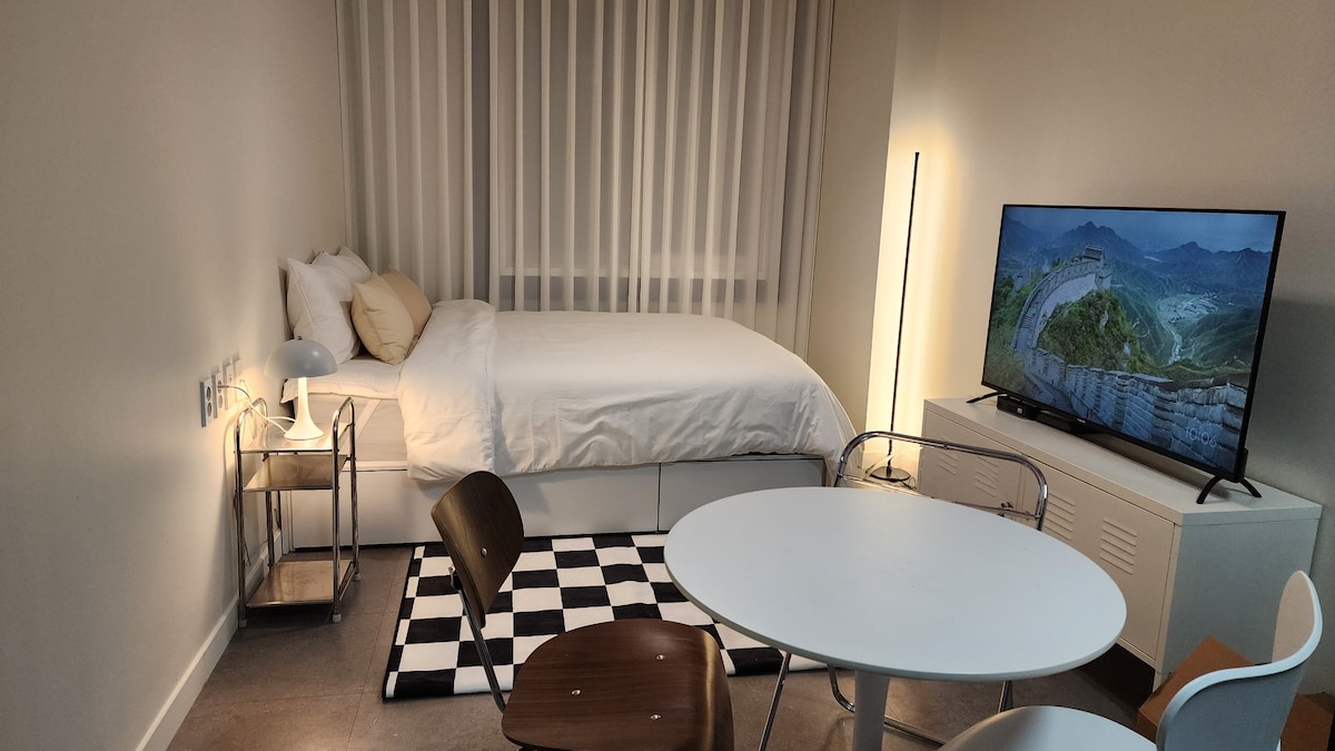 A well-lit room features a spacious bed covered with crisp white linens. A 50-inch smart TV is positioned nearby. A round table with two chairs sits on a patterned rug, while a shelf and a floor lamp add functional elements to the space.