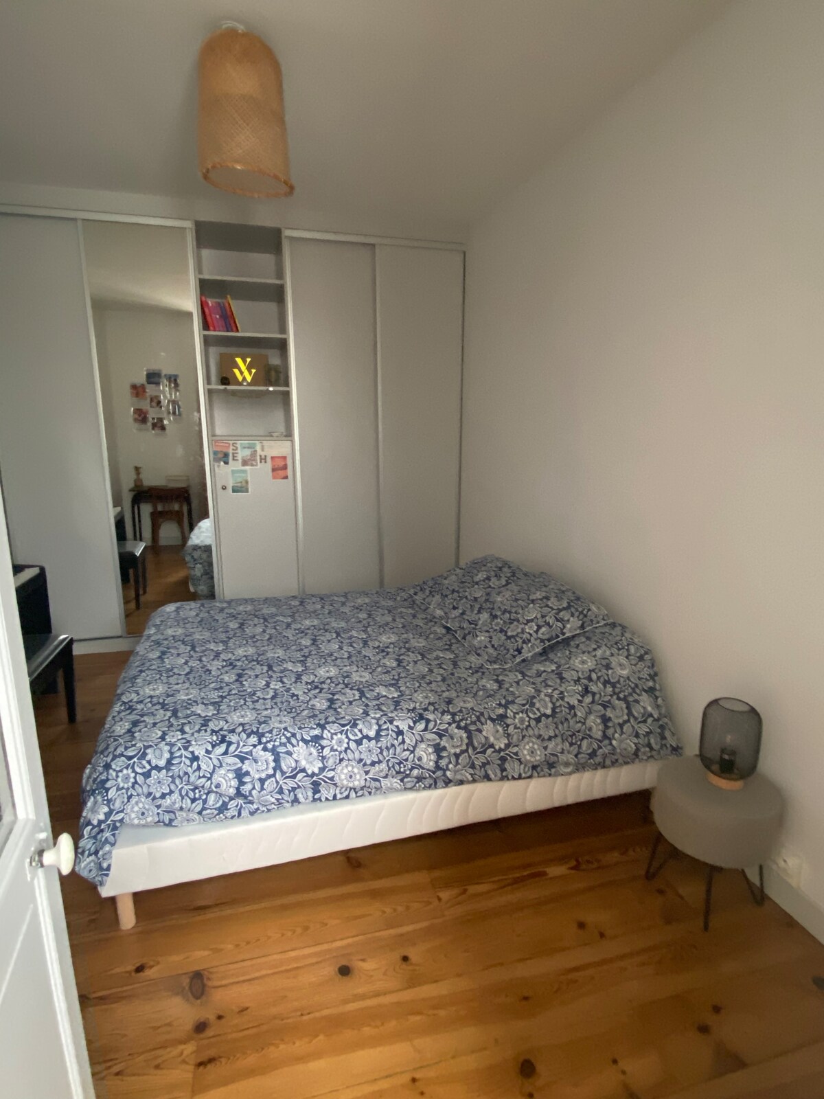 A cozy bedroom is furnished with a spacious bed covered in a blue floral patterned duvet. A small bedside table with a lamp provides soft lighting. Mirrored sliding doors reflect light and create a sense of openness, while wooden flooring adds warmth to the space.