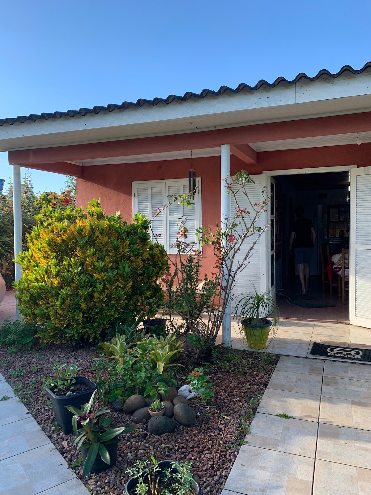 The exterior of a charming house is shown, featuring a welcoming entrance flanked by lush greenery. White shutters are open, showcasing the inviting interior. A pathway lined with decorative plants leads to the entrance, enhancing the home's friendly atmosphere.
