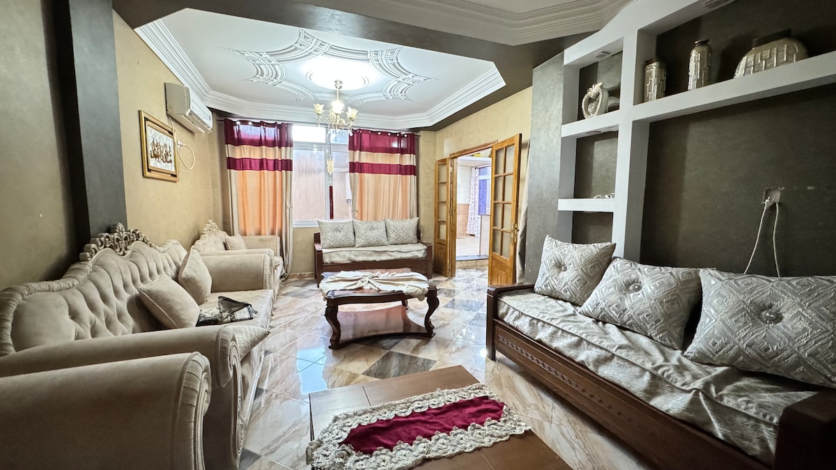 A spacious living area is furnished with elegant sofas and decorative cushions. Natural light enters through large windows covered with light curtains, highlighting the ornate ceiling design. A central coffee table sits on polished marble flooring, complemented by tasteful wall decor.