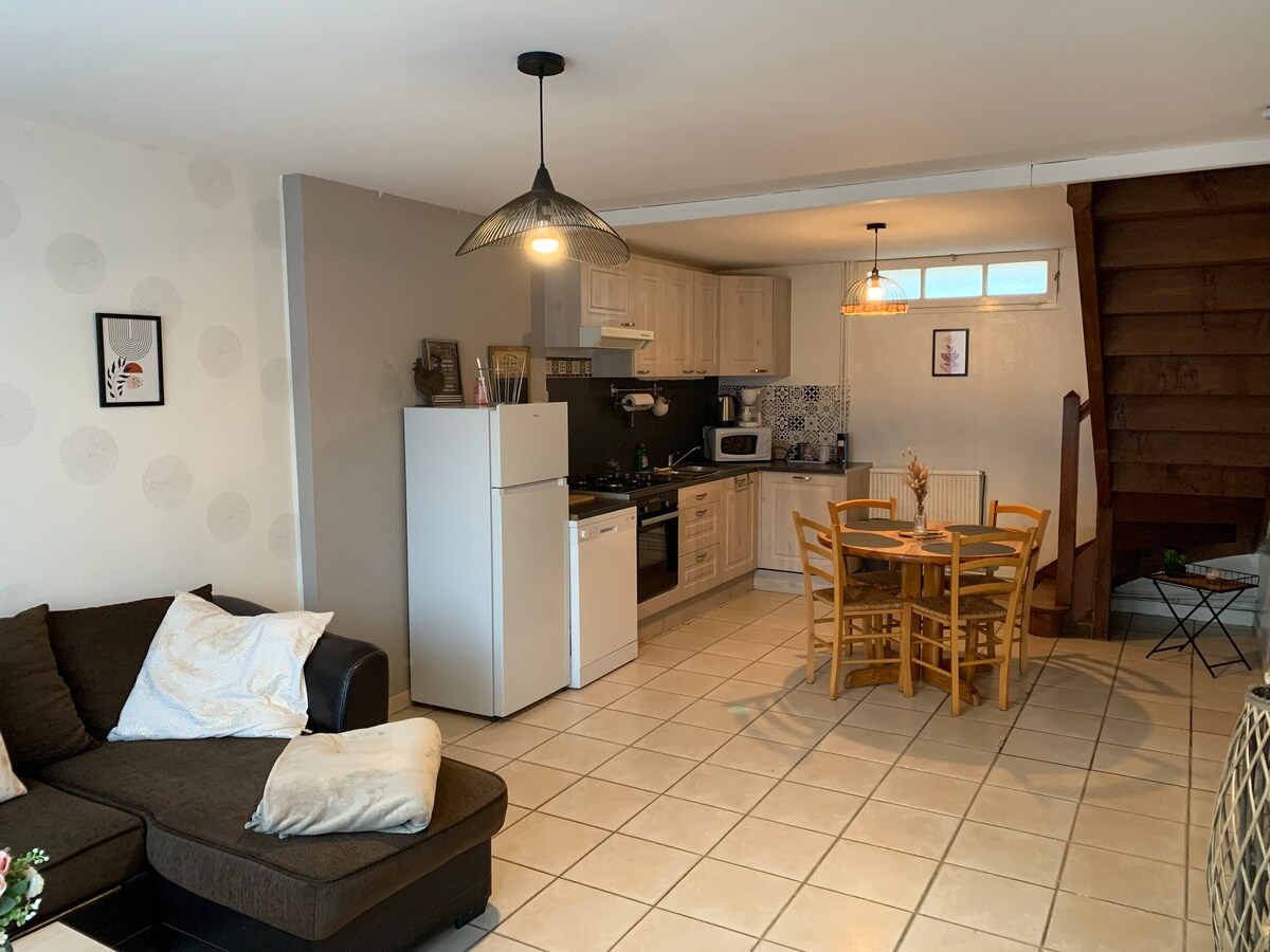 A functional living space features an open kitchen with light cabinetry and essential appliances. A dining table for four sits near the kitchen, surrounded by wooden chairs. A comfortable sectional sofa is positioned in the corner, providing a relaxed seating area. Bright tiles enhance the room's spacious feel.
