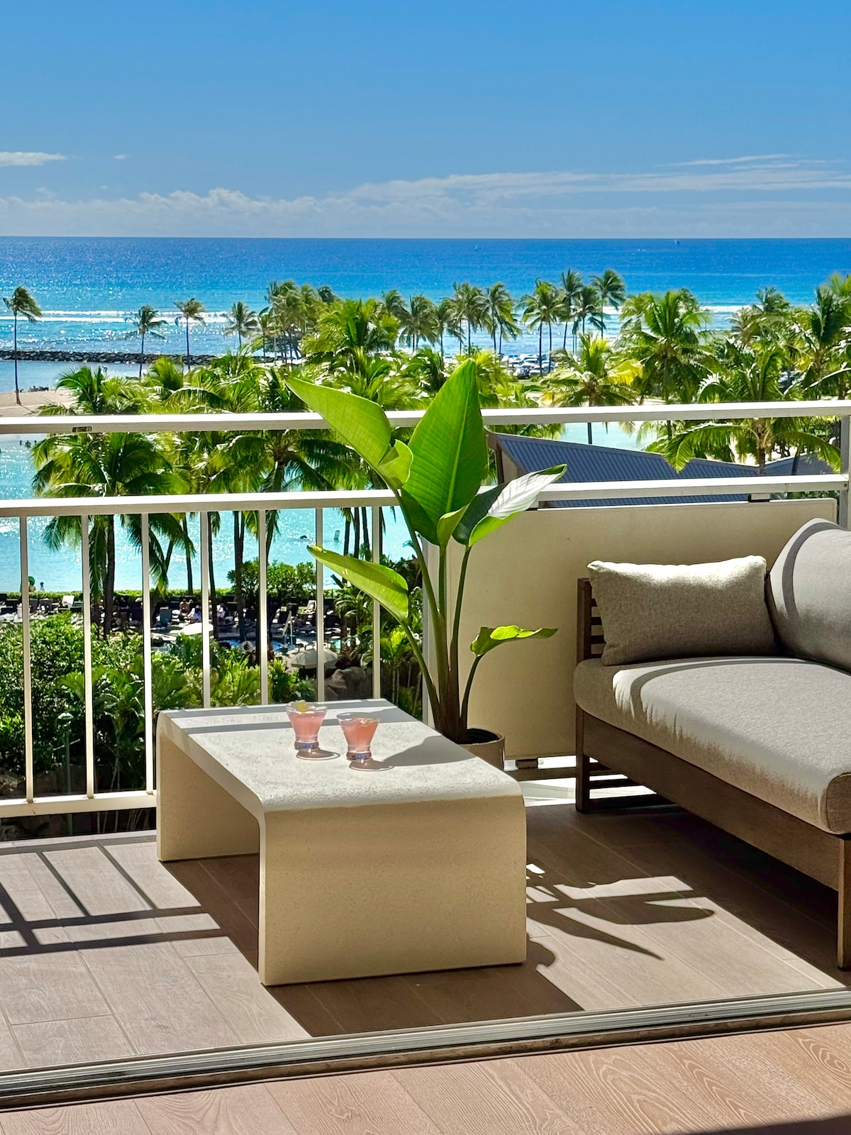 An outdoor balcony space is displayed, featuring a light-colored table with two drinks positioned on it. A cozy sofa with neutral cushions sits against the railing, overlooking the vibrant ocean and palm trees under a clear blue sky.
