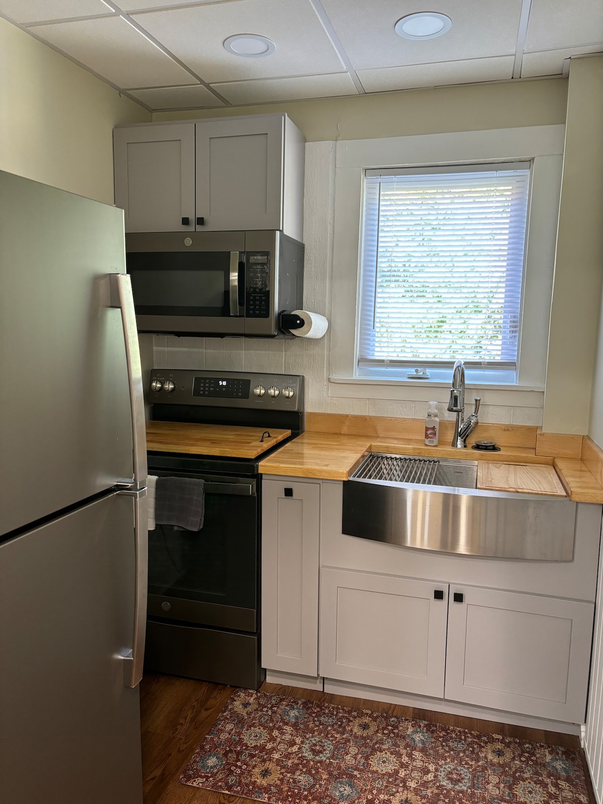 A compact kitchen area is equipped with sleek stainless steel appliances, including a refrigerator and microwave. A modern stove and a stainless steel sink with a polished countertop are featured. Natural light filters through the window above the sink, illuminating the space.