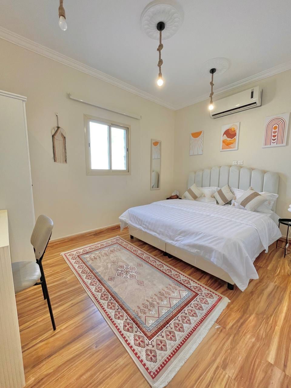 A spacious bedroom features a light-colored bed with decorative pillows, complemented by wooden flooring. Artwork hangs on the wall above the bed, while natural light enters through a window. A small table and chair are placed to the side, enhancing the room's functionality.