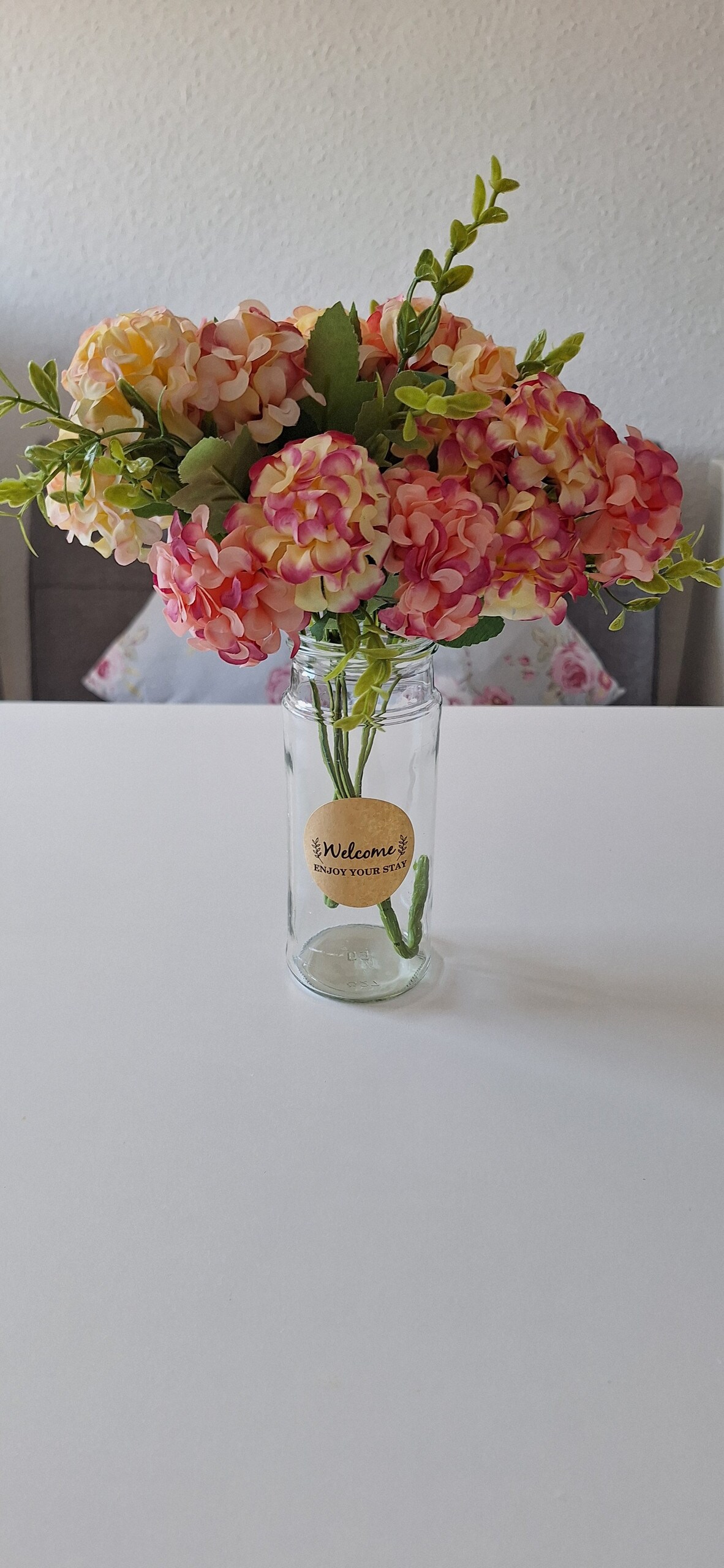 A clear glass jar holds an arrangement of artificial flowers in shades of pink and peach. The jar features a label that reads 'Welcome!' in a friendly font, while the table provides a simple, clean backdrop, enhancing the floral display.