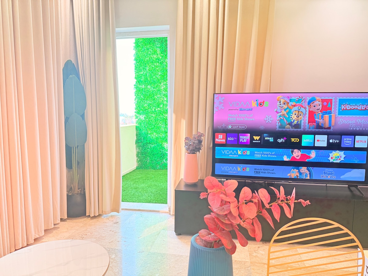 A living space features a large TV displaying colorful content. Flowing curtains frame a sliding door that opens to a green outdoor area. A round table with a light tablecloth and a decorative plant adds a touch of color to the room.