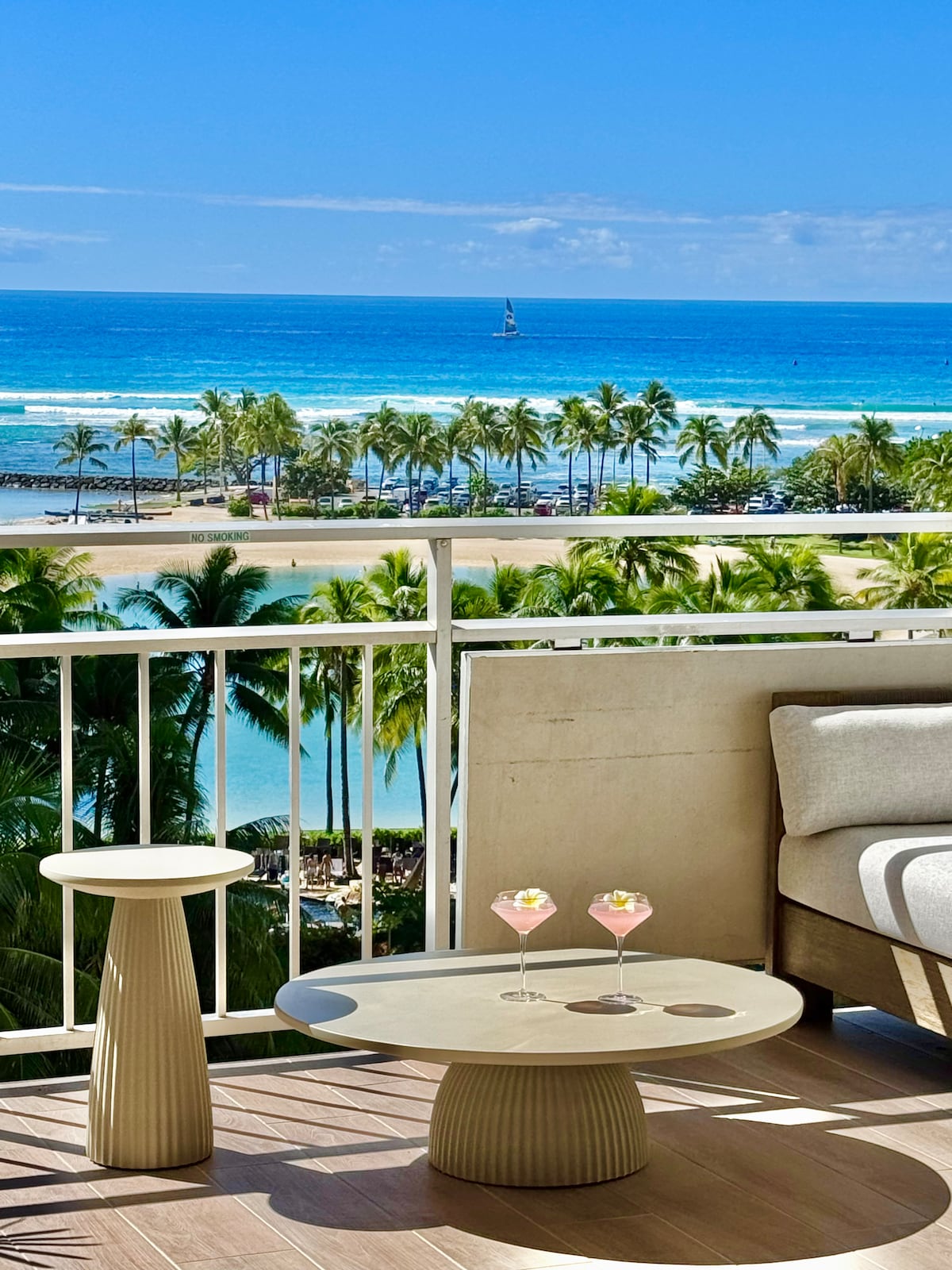 A spacious balcony offers a direct view of the ocean and beach. Two drinks are placed on a round table, accompanied by a comfortable outdoor sofa. Lush palm trees line the beach, enhancing the serene coastal scenery.