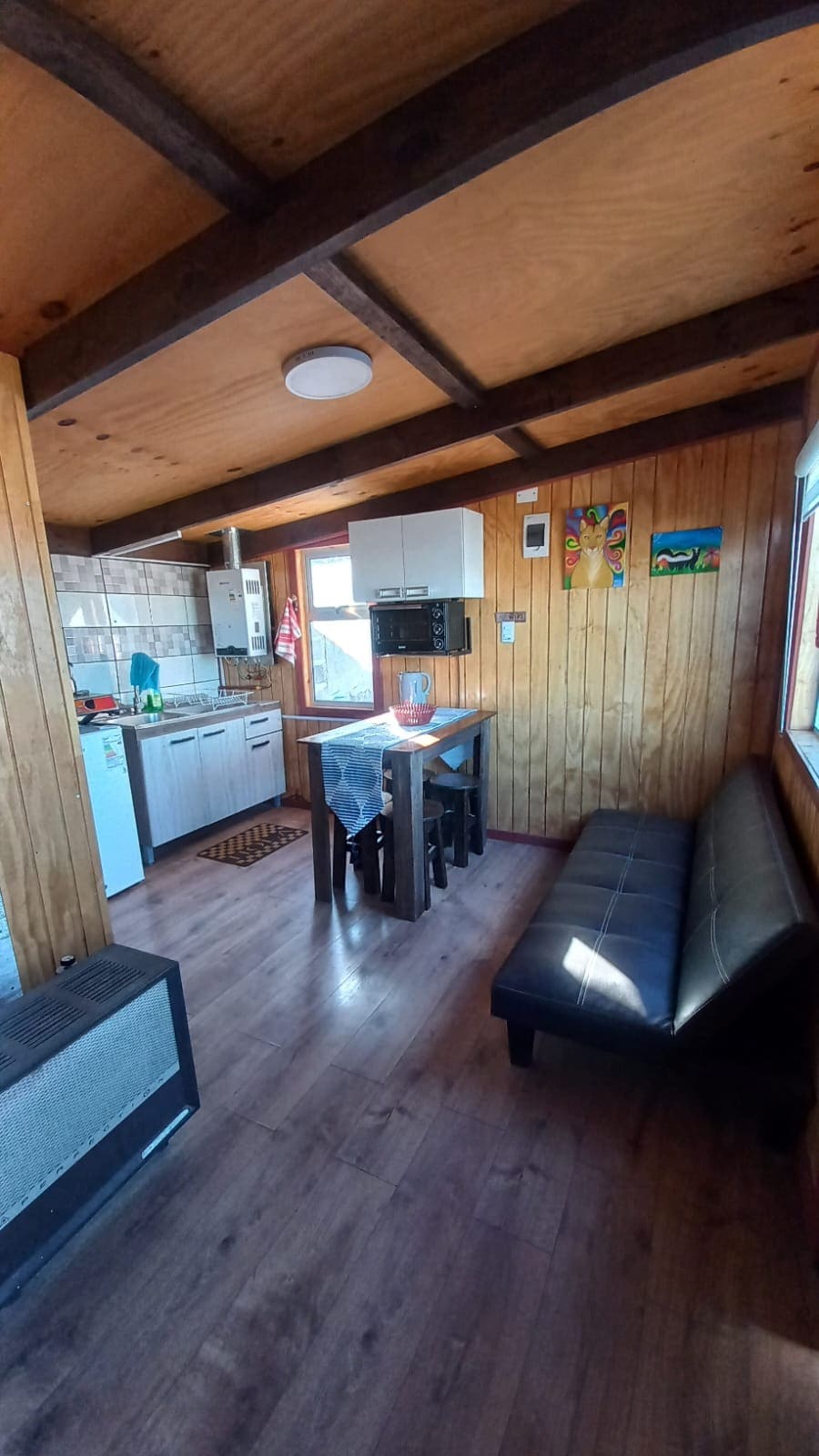 An open living space showcases a compact kitchen with modern appliances and wooden cabinetry. A dining table is set for four, while a dark sofa adds comfort to the area. Natural light fills the room through multiple windows, enhancing the warm wooden interior.