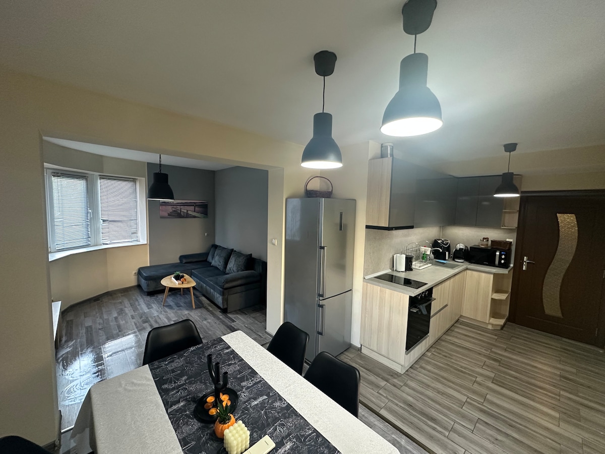 An open-plan kitchen and dining area features modern appliances and a light-colored countertop. A dining table with dark chairs is positioned centrally, while a cozy living area with a comfortable sofa is visible in the background. Warm lighting enhances the spacious layout.