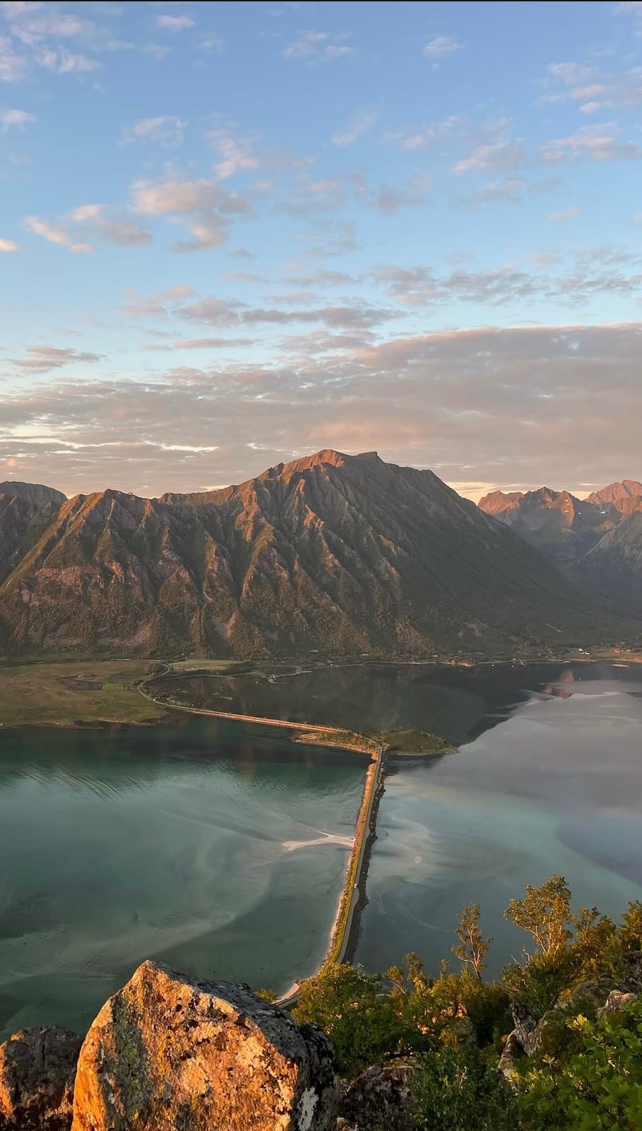 A scenic view is captured, showcasing a majestic mountain range with rugged peaks reflecting in a calm body of water below. A winding road stretches along the coastline, bordered by lush green vegetation, under a sky painted with soft hues of orange and blue.