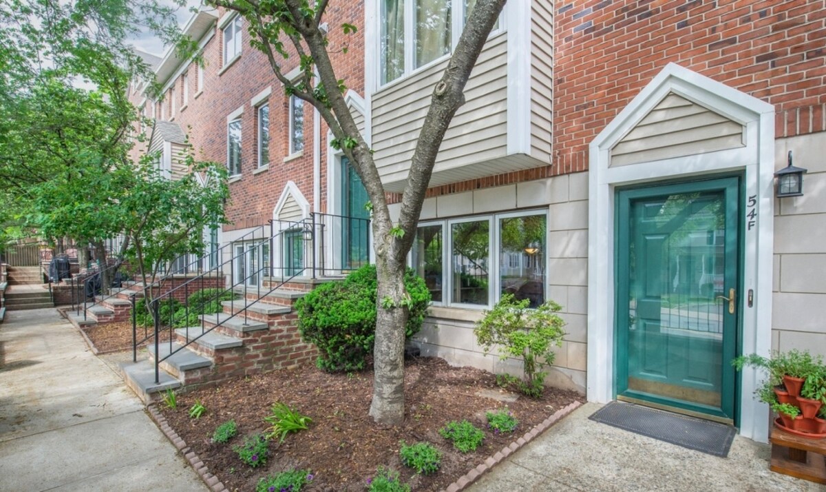 A welcoming entrance features a green front door, framed by light-colored siding and brick exterior. Lush greenery and flower beds create a pleasant atmosphere. A set of steps leads to the entrance, while large windows allow natural light to illuminate the space.