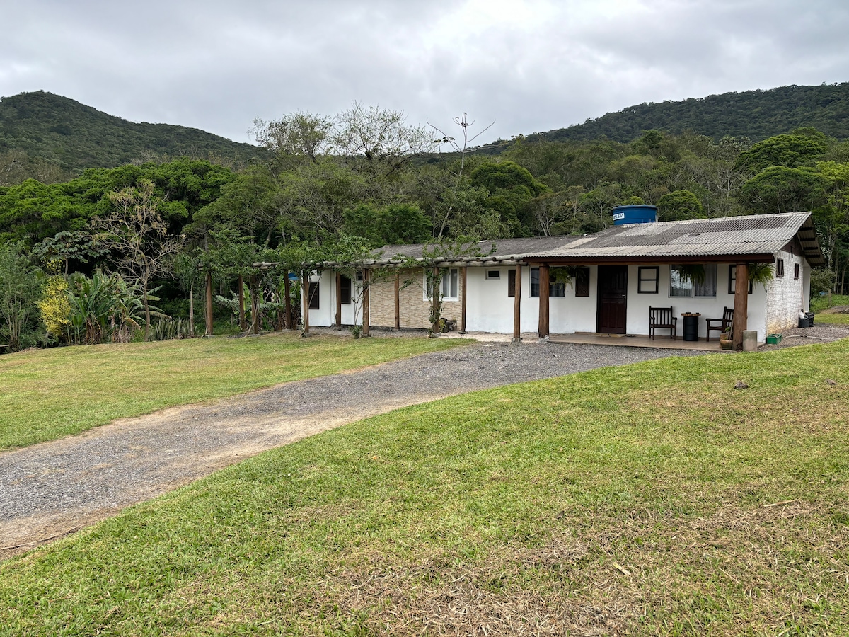 A rustic hostel is situated among lush greenery, with a gravel pathway leading to its entrance. The building features a mix of natural stone and white walls, complemented by a sloped metal roof. Surrounding hills add to the serene, natural environment.