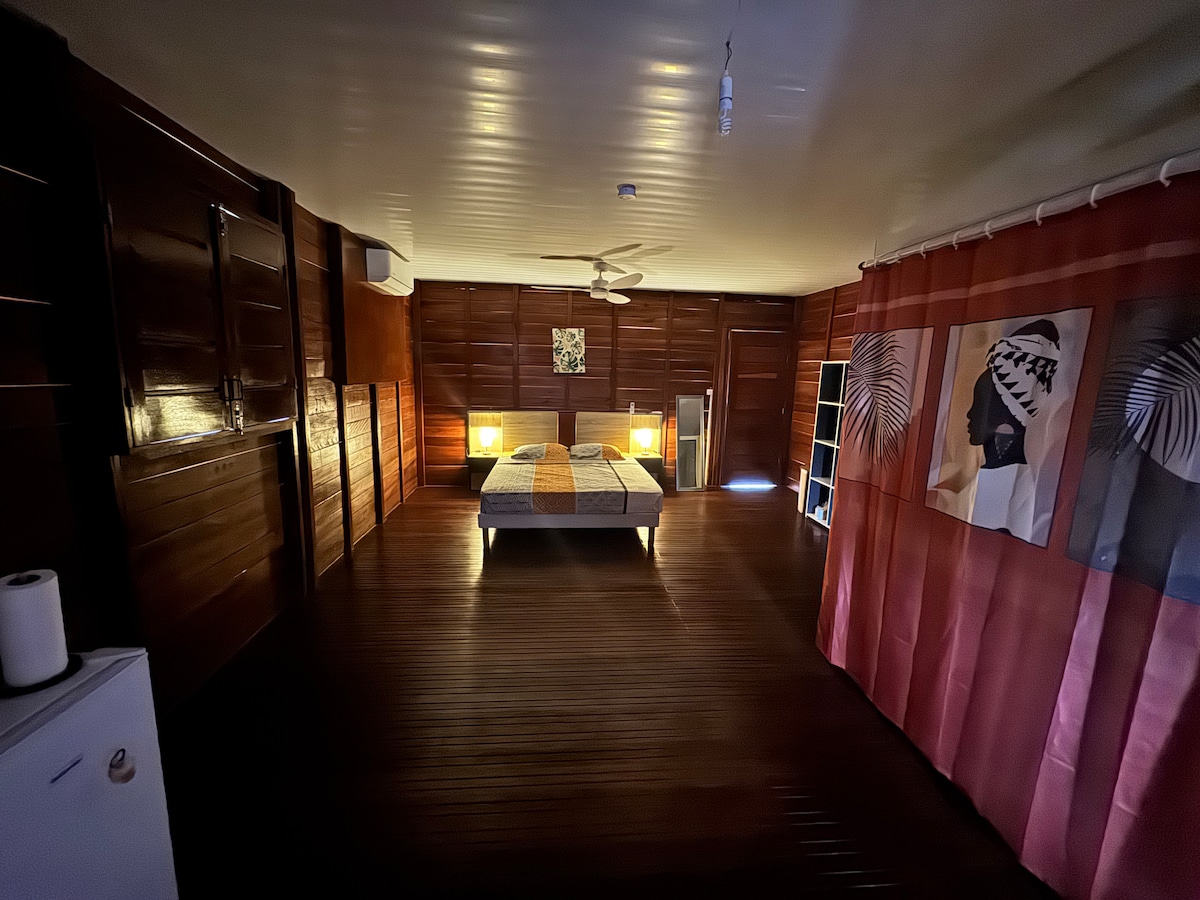 A spacious room features dark wooden paneling and a large bed positioned at the center. Soft lighting from bedside lamps creates a warm ambiance. A small refrigerator and a bookshelf are visible, alongside two decorative art pieces on the wall, enhancing the tranquil setting.