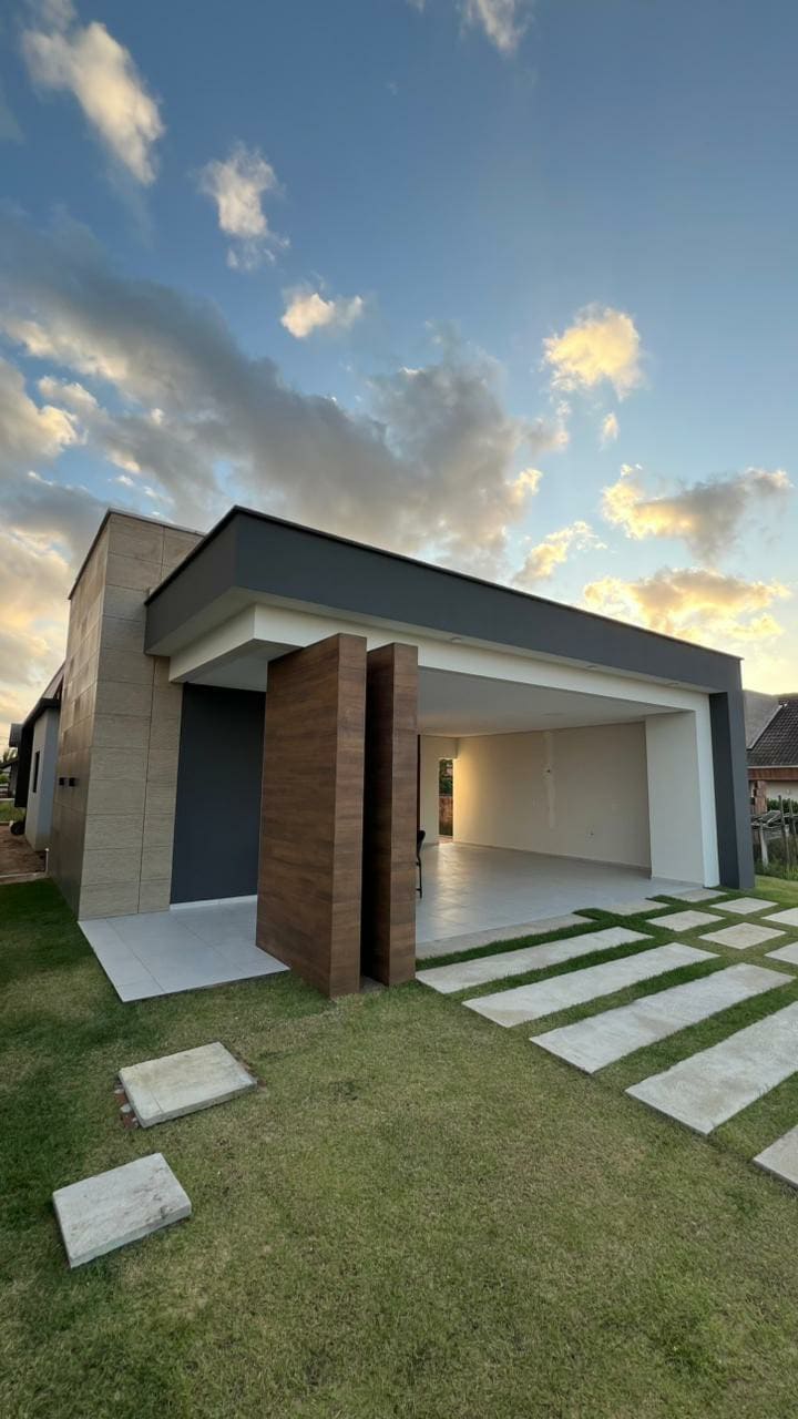 The image depicts a modern house with a striking entrance featuring large wooden doors. A spacious driveway leads to the front, complemented by neatly arranged grass and stone pathways. The sky is filled with clouds, hinting at a serene atmosphere.