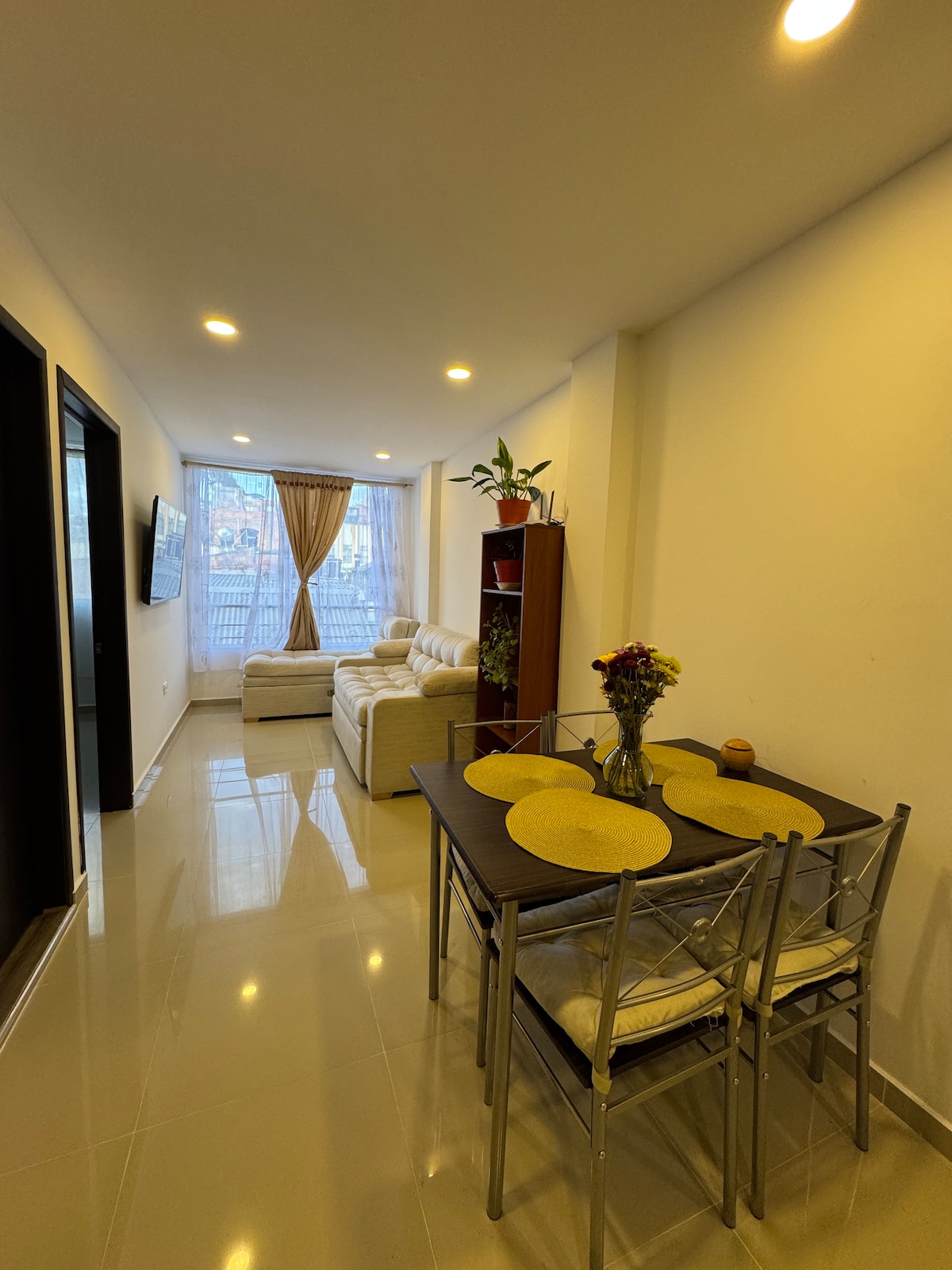 A bright living space features a light-colored sofa positioned near a window with sheer curtains. A small dining table set for four is displayed, accompanied by a decorative vase of flowers. Warm lighting highlights the glossy tile floor, adding to the overall spacious feel.