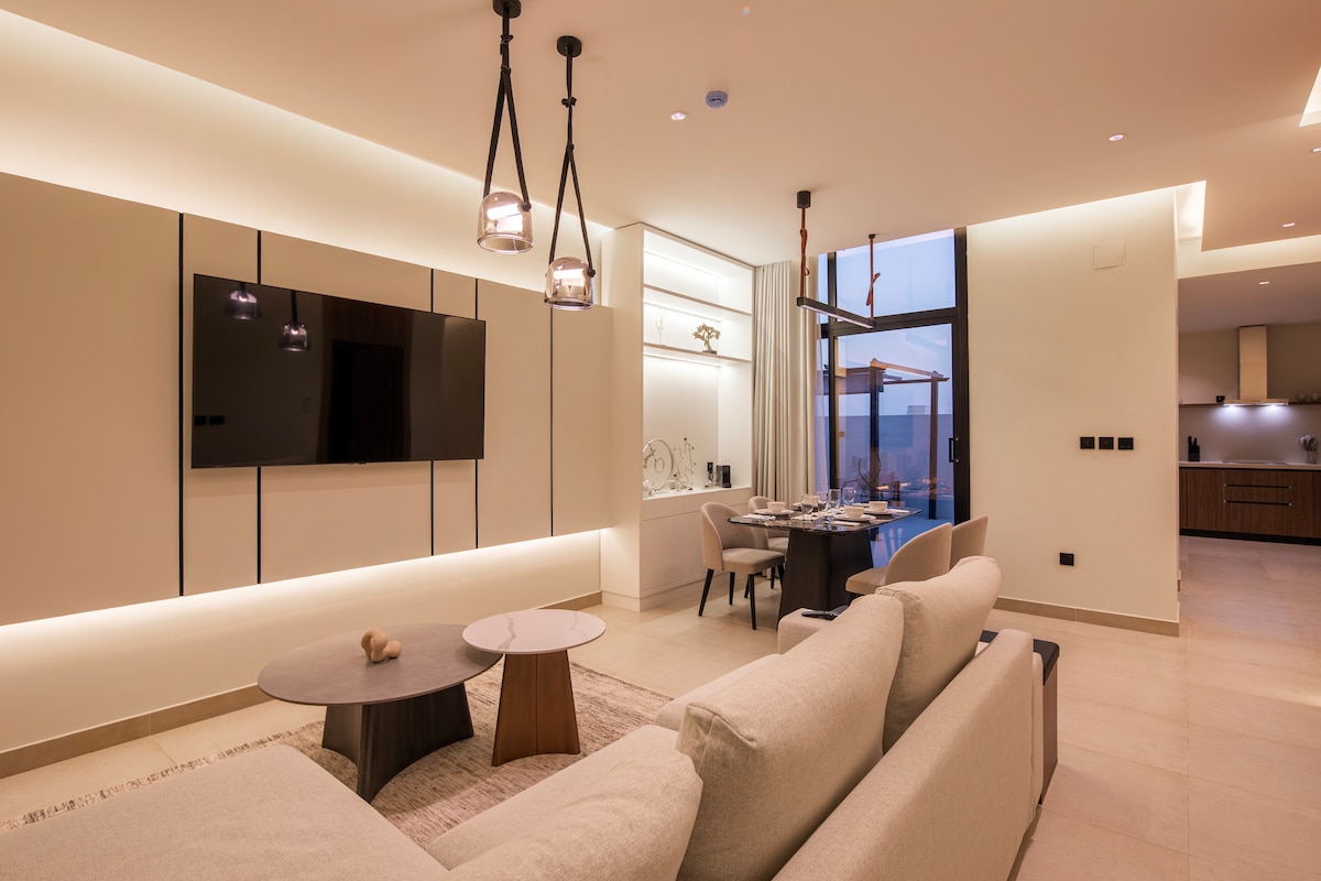 A contemporary living space features a plush sofa and stylish coffee tables. A wall-mounted TV is positioned above an elegant sideboard. Natural light filters through expansive windows, illuminating the dining area set for guests, with a view of the surrounding area.