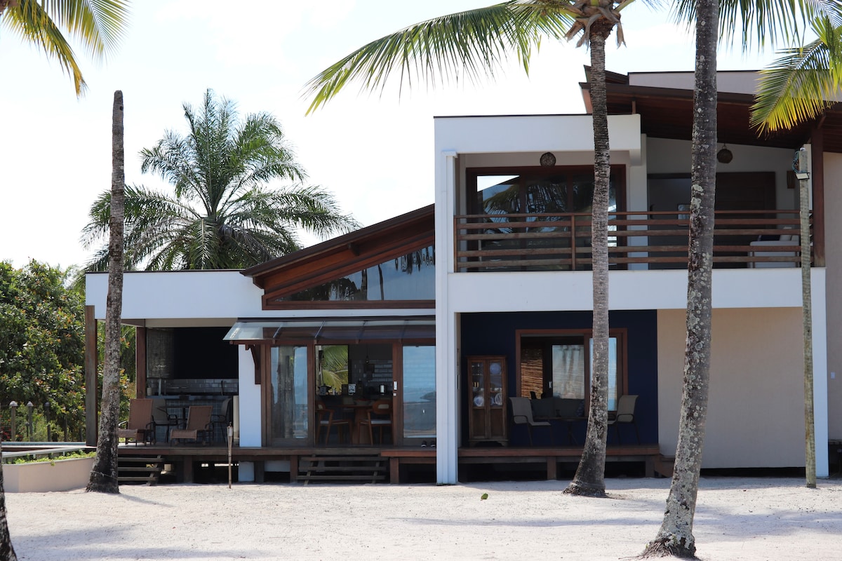 A modern coastal house is set amidst palm trees, showcasing a blend of natural materials and clean lines. Large windows offer views of the surrounding greenery, while an outdoor patio with seating is accessible from the main entrance.