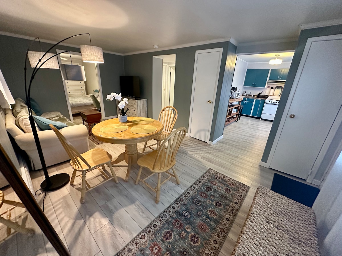 A cozy living area features a round wooden table with four chairs, accompanied by a floor lamp with two shades. A beige sofa is positioned next to a large mirror, with a glimpse of the kitchen visible in the background, showcasing blue cabinets and modern appliances.