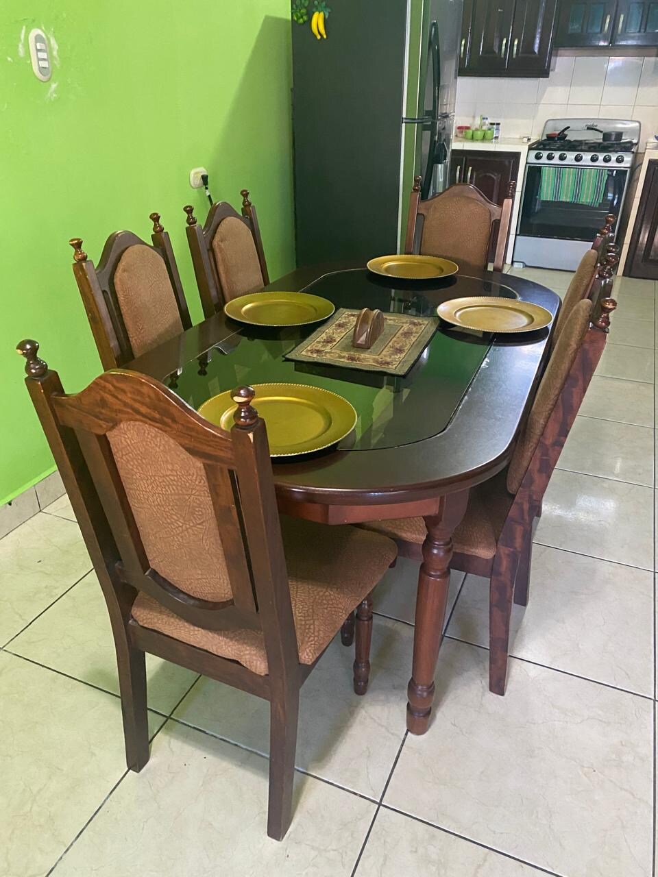 A wooden dining table is surrounded by six upholstered chairs. Plates are arranged on the table's surface, complemented by a decorative centerpiece. The vibrant green walls provide a bright backdrop, and kitchen appliances are visible in the background.