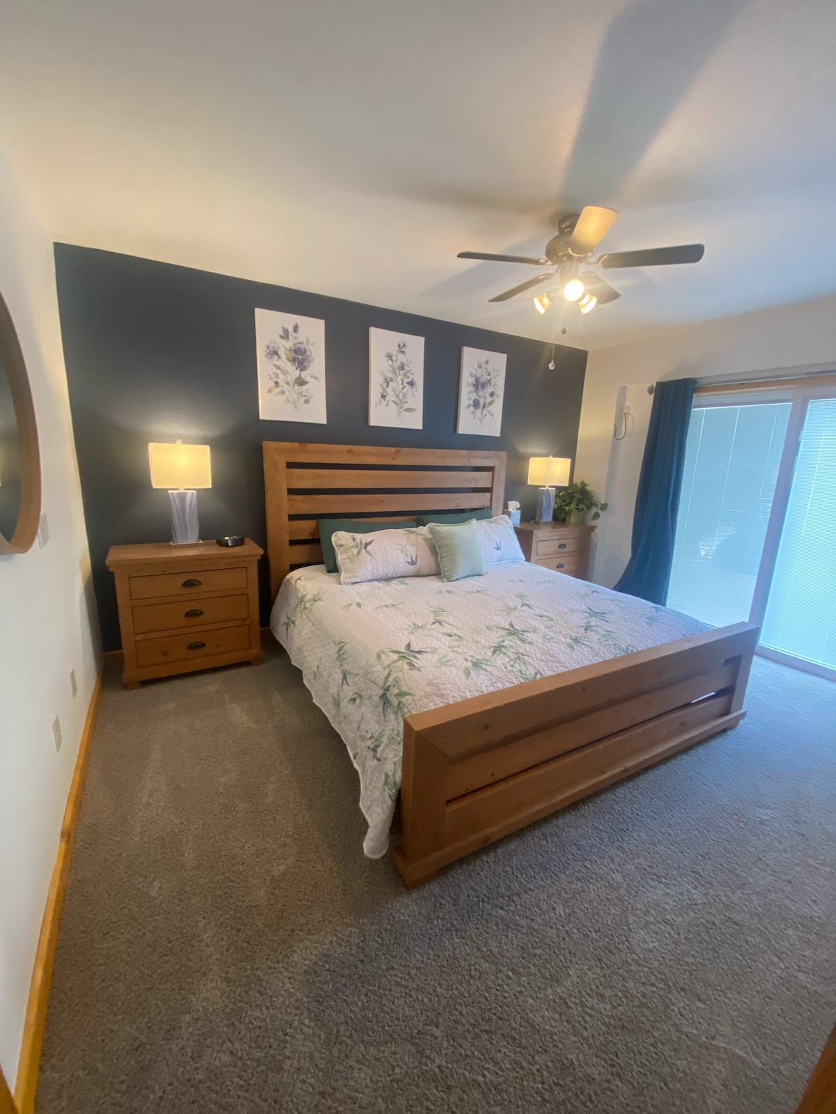 A cozy bedroom features a wooden bed frame with a floral-patterned comforter. Two lamps on nightstands provide soft lighting, while a large window with sheer curtains allows natural light to enter. Floral wall art is displayed above the bed, complementing the neutral wall.