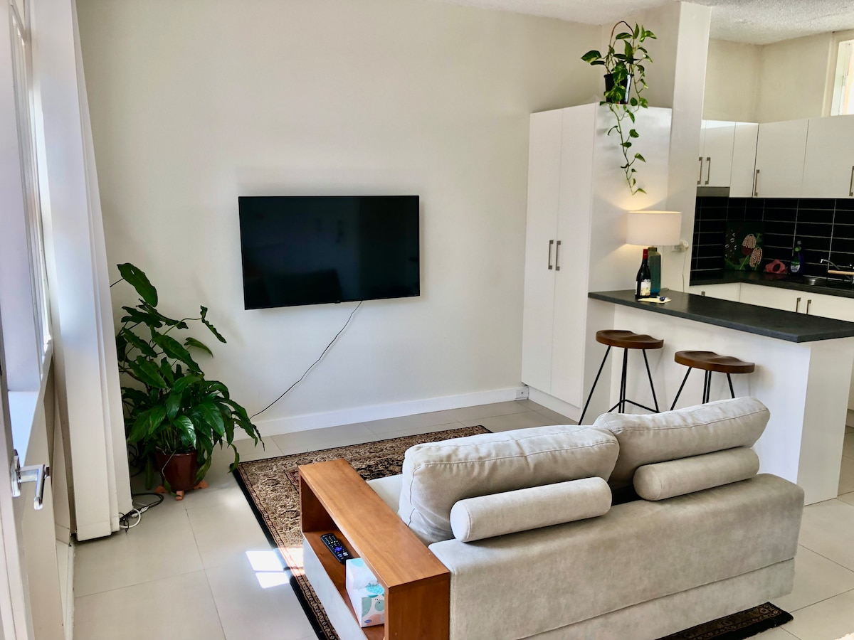 A modern living area is showcased with a sectional sofa facing a wall-mounted TV. A small wooden coffee table rests on a patterned rug. Bar stools are positioned near a sleek kitchen with white cabinetry and dark countertops, while a leafy plant adds a touch of greenery.