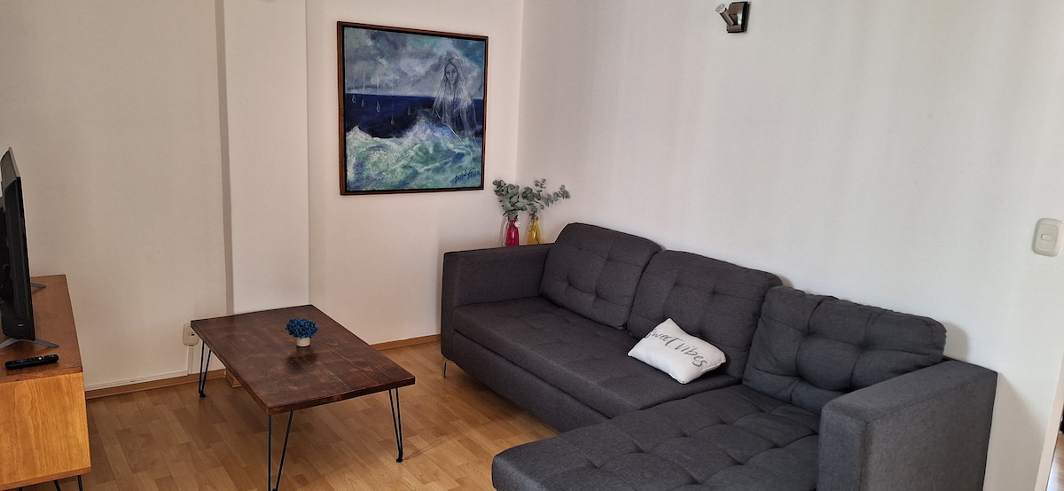 A living area features a modern gray sectional sofa alongside a wooden coffee table. A large wall art piece depicting a seascape hangs above the couch. A small blue decorative item is placed on the table, while natural light brightens the space.
