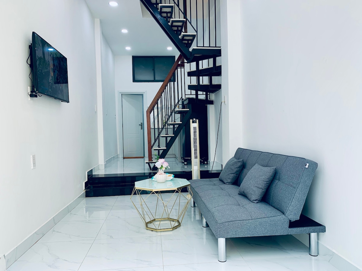 A modern living area is seen, featuring a gray futon and a decorative gold coffee table with delicate floral arrangements. A wall-mounted television is visible, and a staircase leads to the upper level. Bright lights illuminate the spacious, tiled floor.